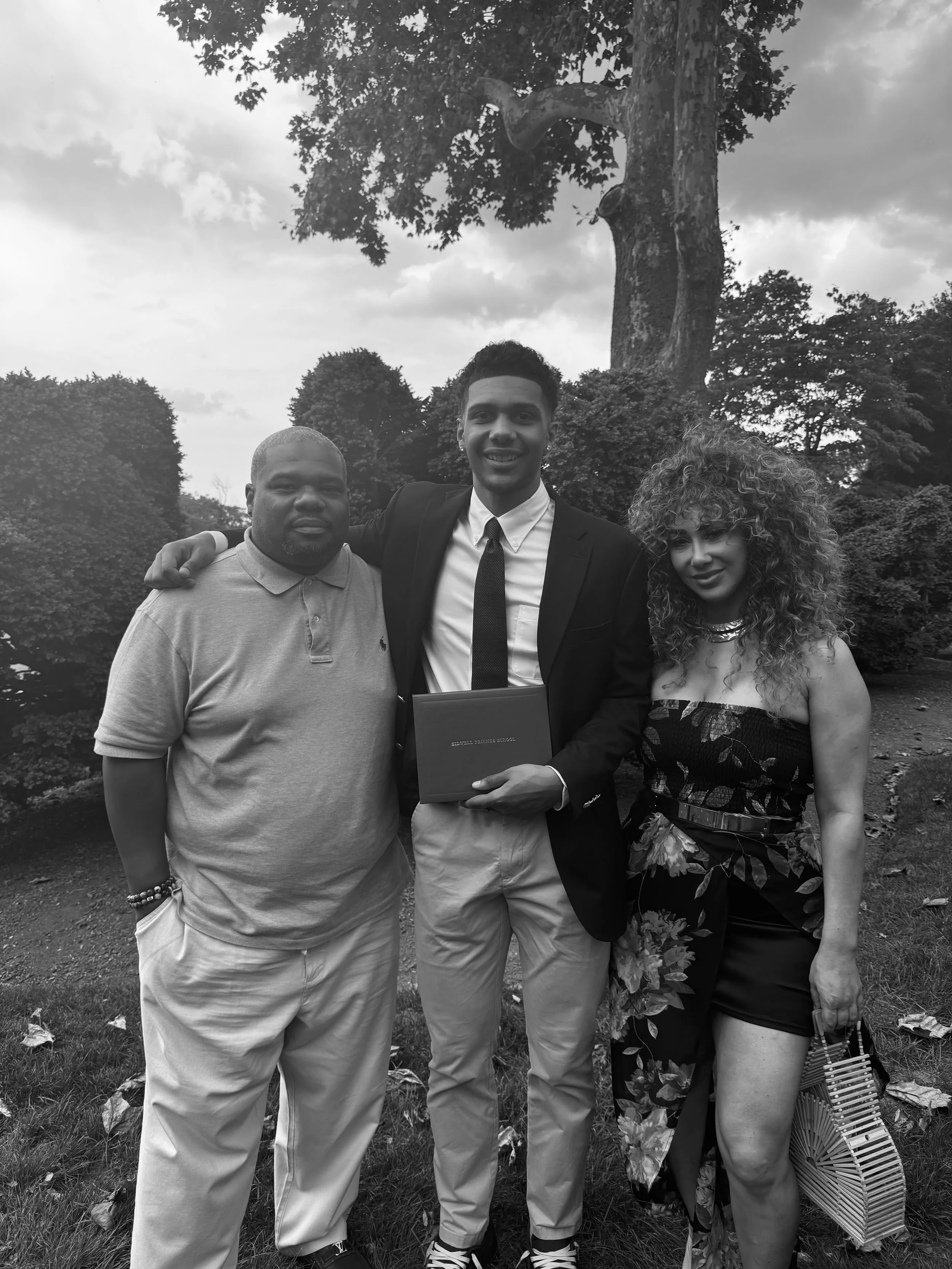 Three people standing outdoors in front of a large tree, dressed in semi-formal and casual attire, smiling at the camera.