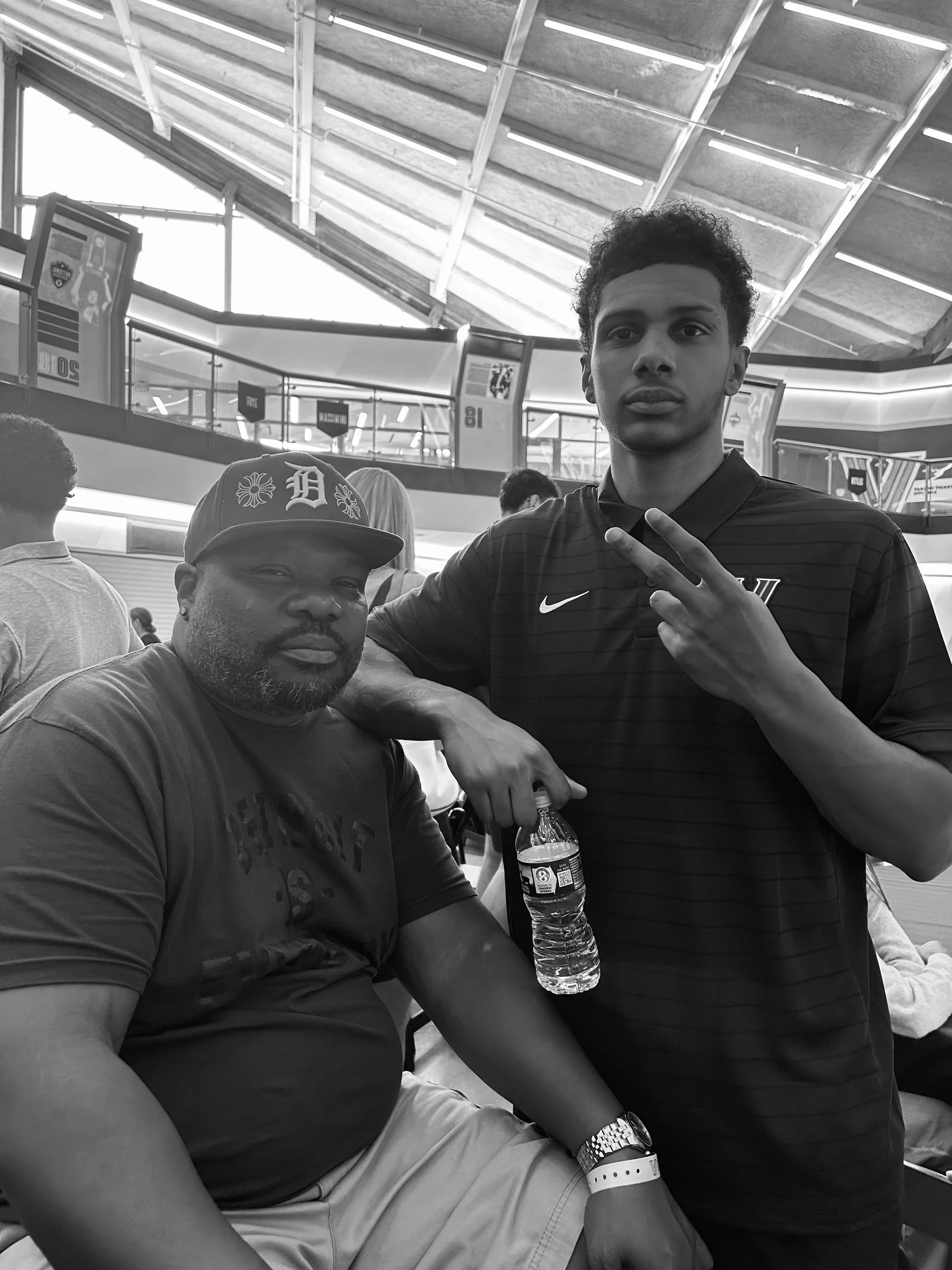 Two men posing for a photo inside a large indoor sports arena. One man is sitting, wearing a baseball cap and a t-shirt, holding a water bottle. The other man is standing, making a peace sign with his fingers, dressed in a sports jersey. The arena ha