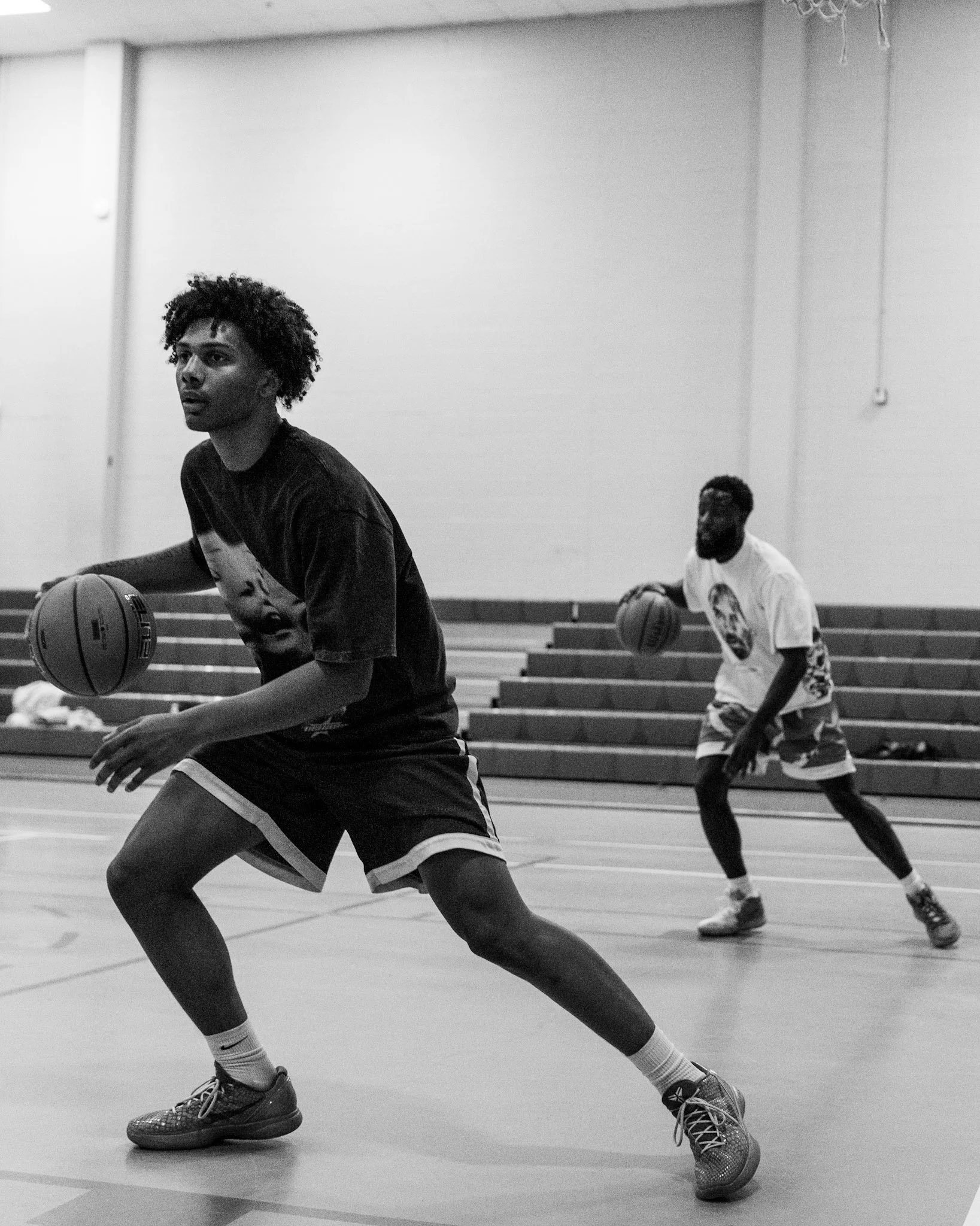 Two young men playing basketball in an indoor gym, one in the foreground dribbling the ball and another in the background preparing to defend.