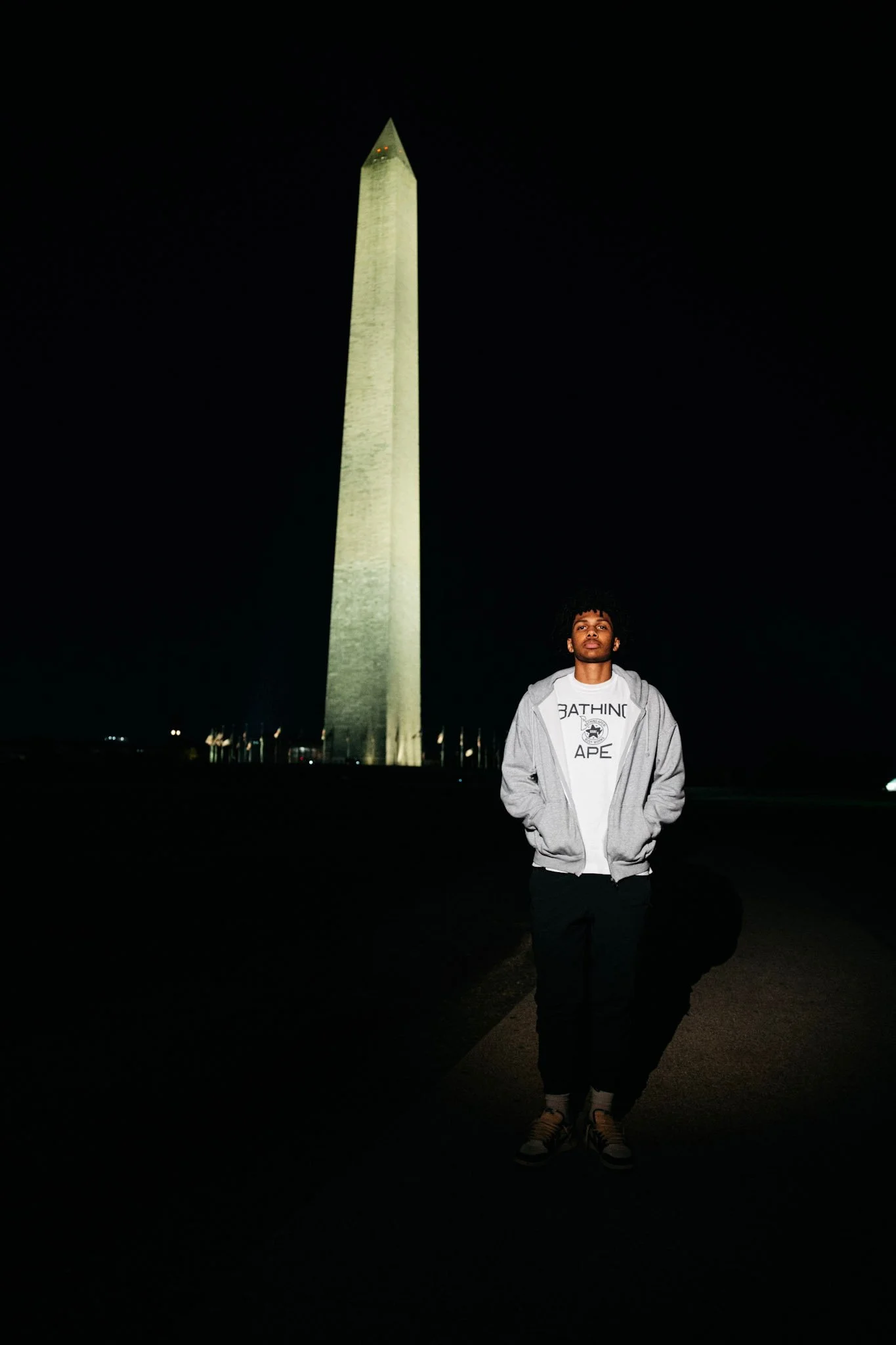 A young man stands in front of the Washington Monument at night, wearing a gray hoodie and a white T-shirt, with his hands in his pockets.
