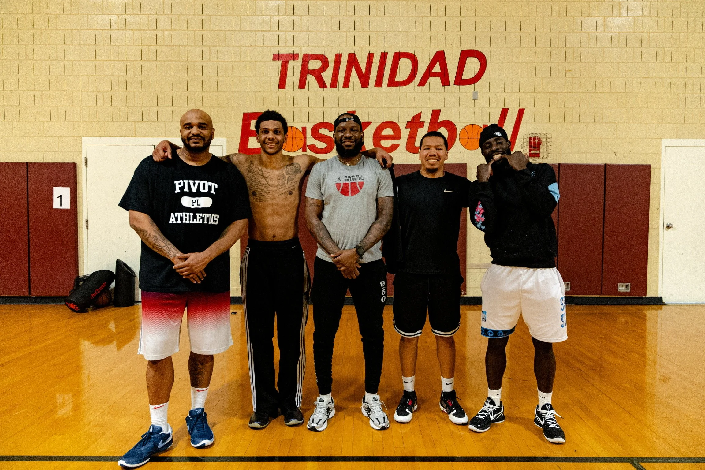 Six men standing together on a basketball court in front of a wall with