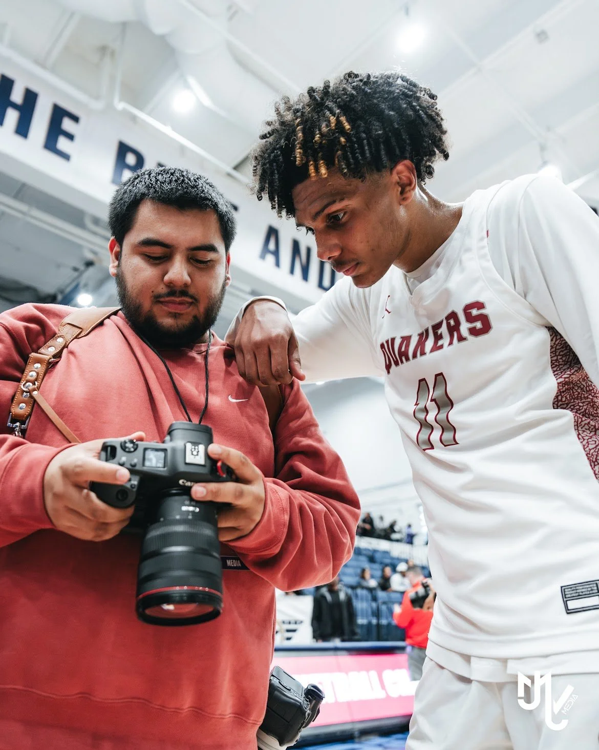 Two men, one in a white sports jersey and the other in a red hoodie, are looking at a camera while one of them rests his arm on the other's shoulder in an indoor sports arena.