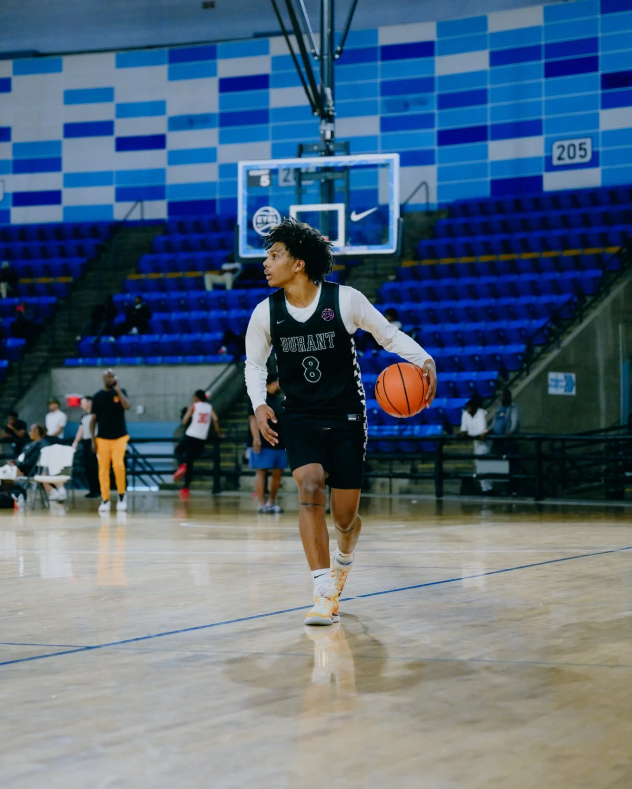A young basketball player in a black and white uniform dribbling a basketball on an indoor court with empty blue bleachers in the background.