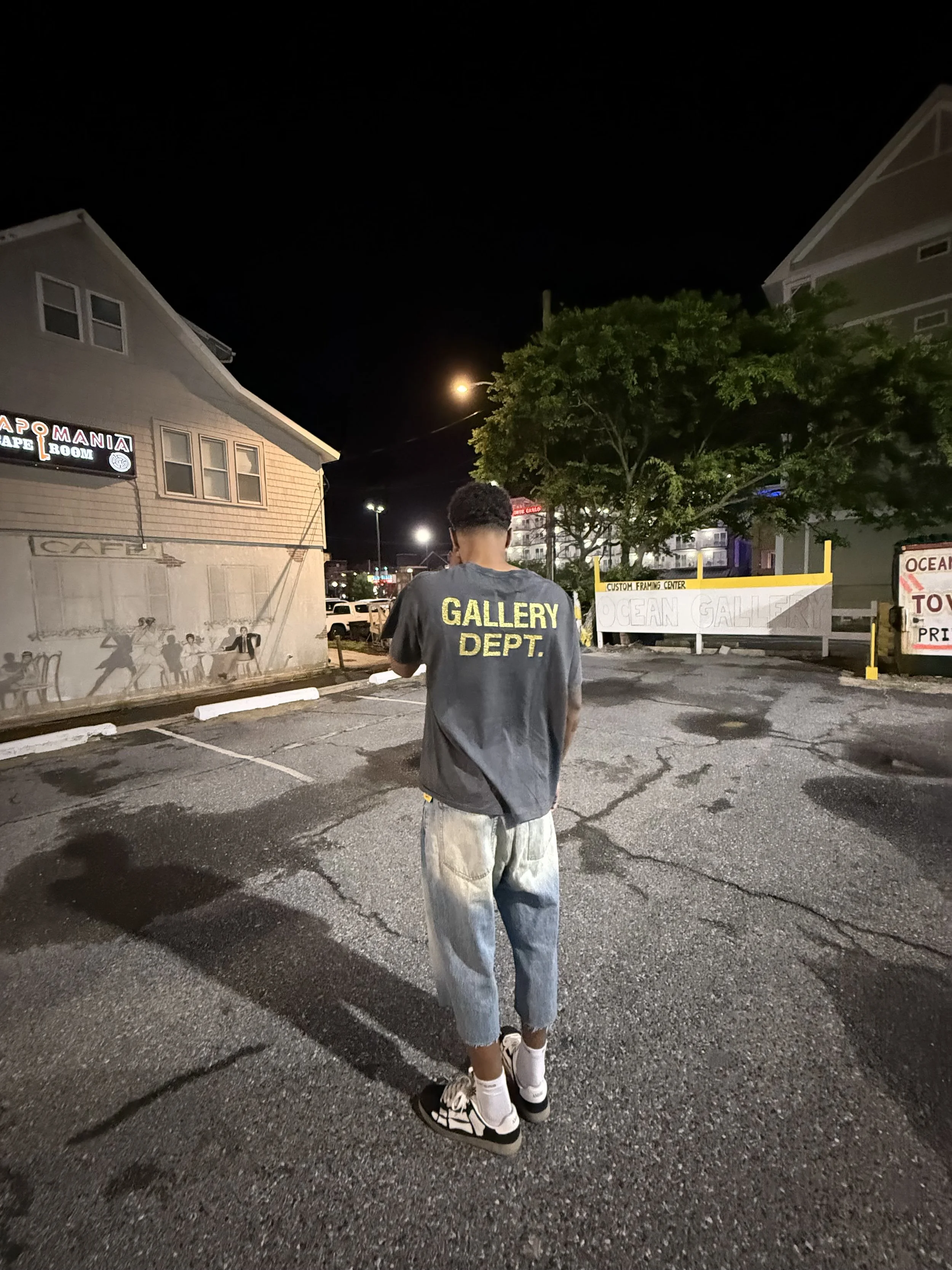 A person wearing a black t-shirt with yellow text that says 'GALLERY DEPT.' standing in an outdoor parking lot at night, facing away from the camera. The parking lot is partially lit, with buildings and signs visible in the background.