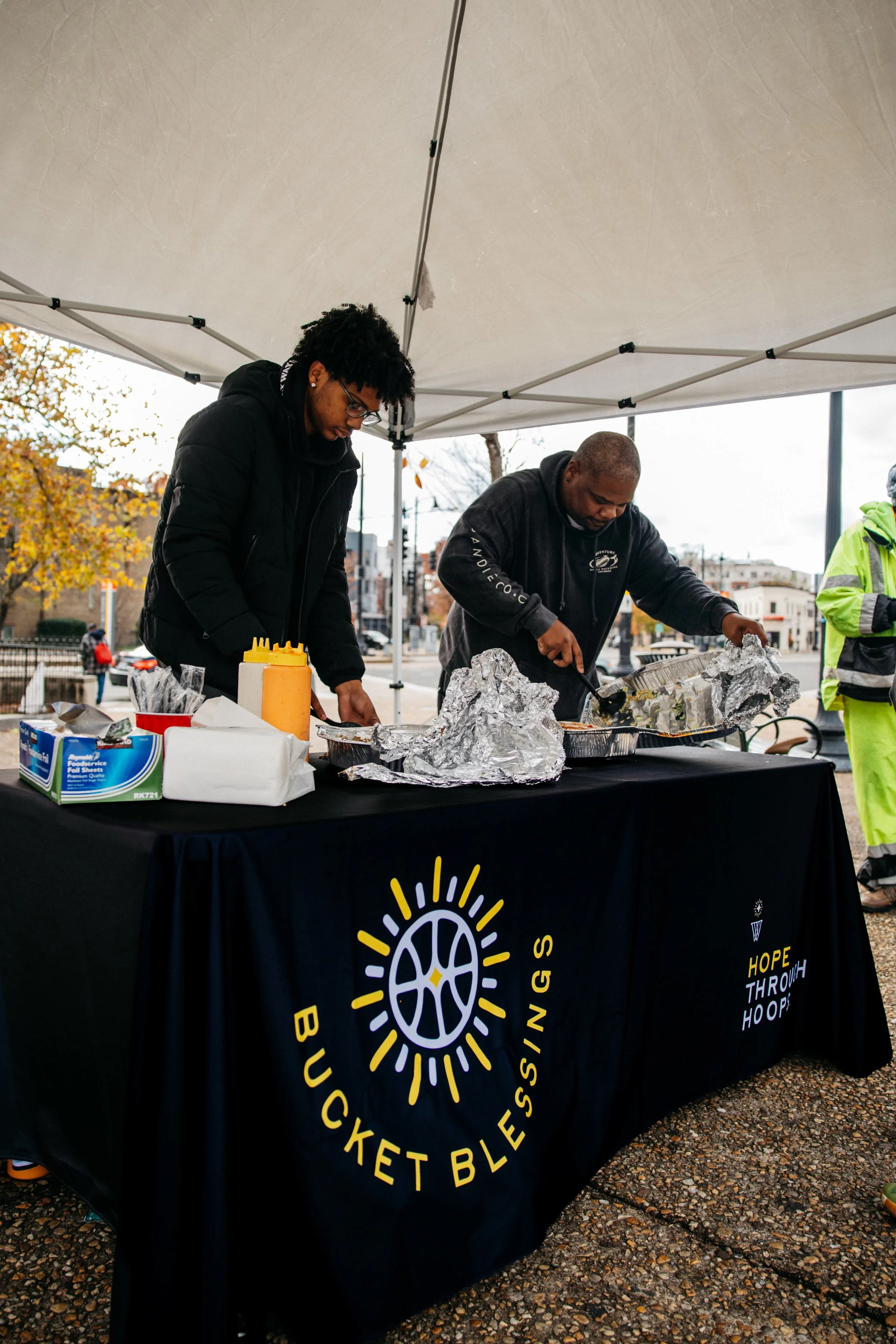 Two people serving food at a table with a Buckett Blessings banner, under a canopy, outdoors with trees and buildings in the background.