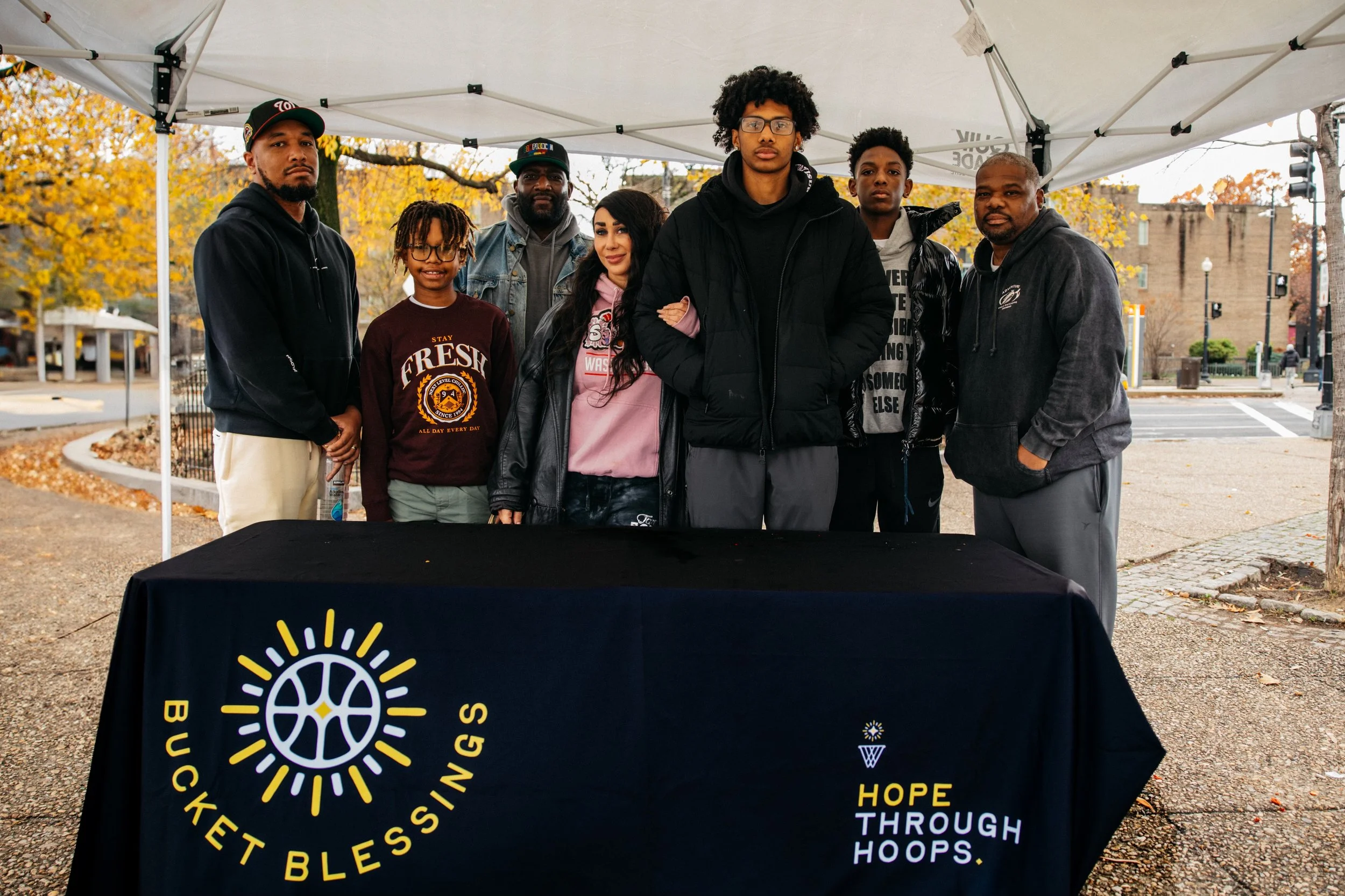 A diverse group of seven people standing behind a table draped with a Buckets Blessings banner, outdoors under a white canopy, with fall trees and a street in the background.