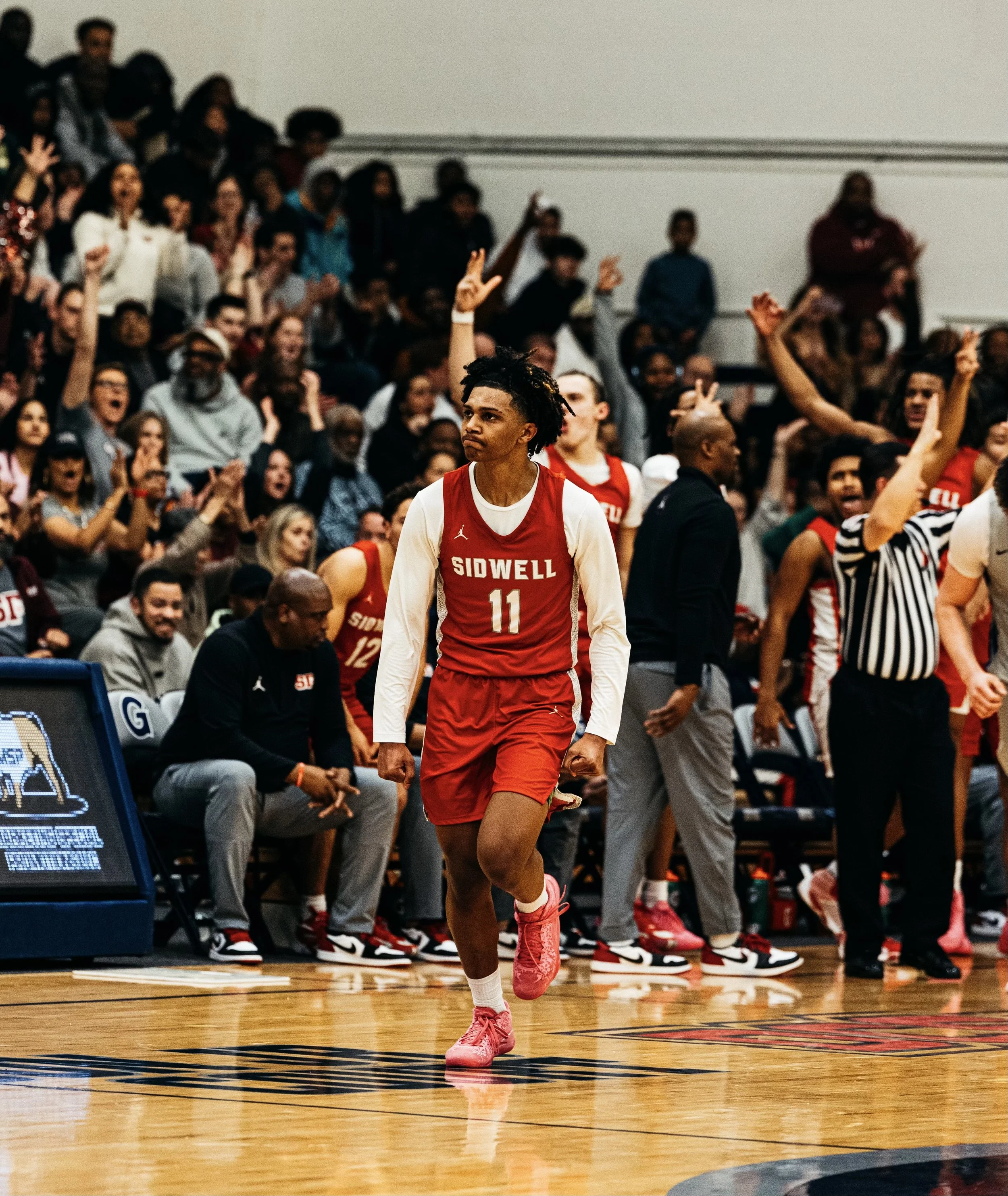 A basketball player wearing a red SIDWELL jersey with the number 11 walks on a basketball court surrounded by cheering teammates and spectators.