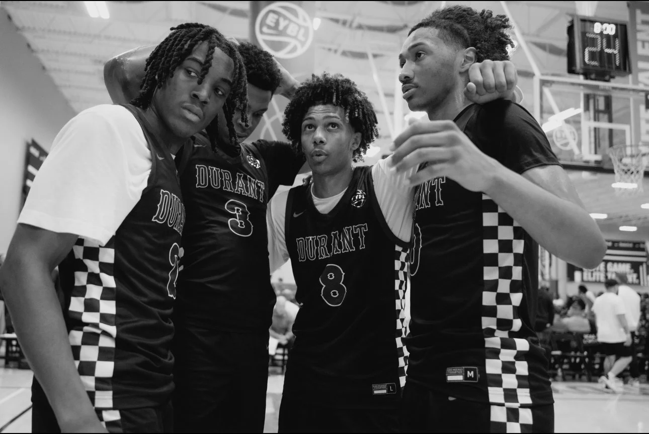 Four basketball players wearing black jerseys with 'DURANT' written on them, standing together in a gymnasium, appearing to discuss strategy or motivate each other.