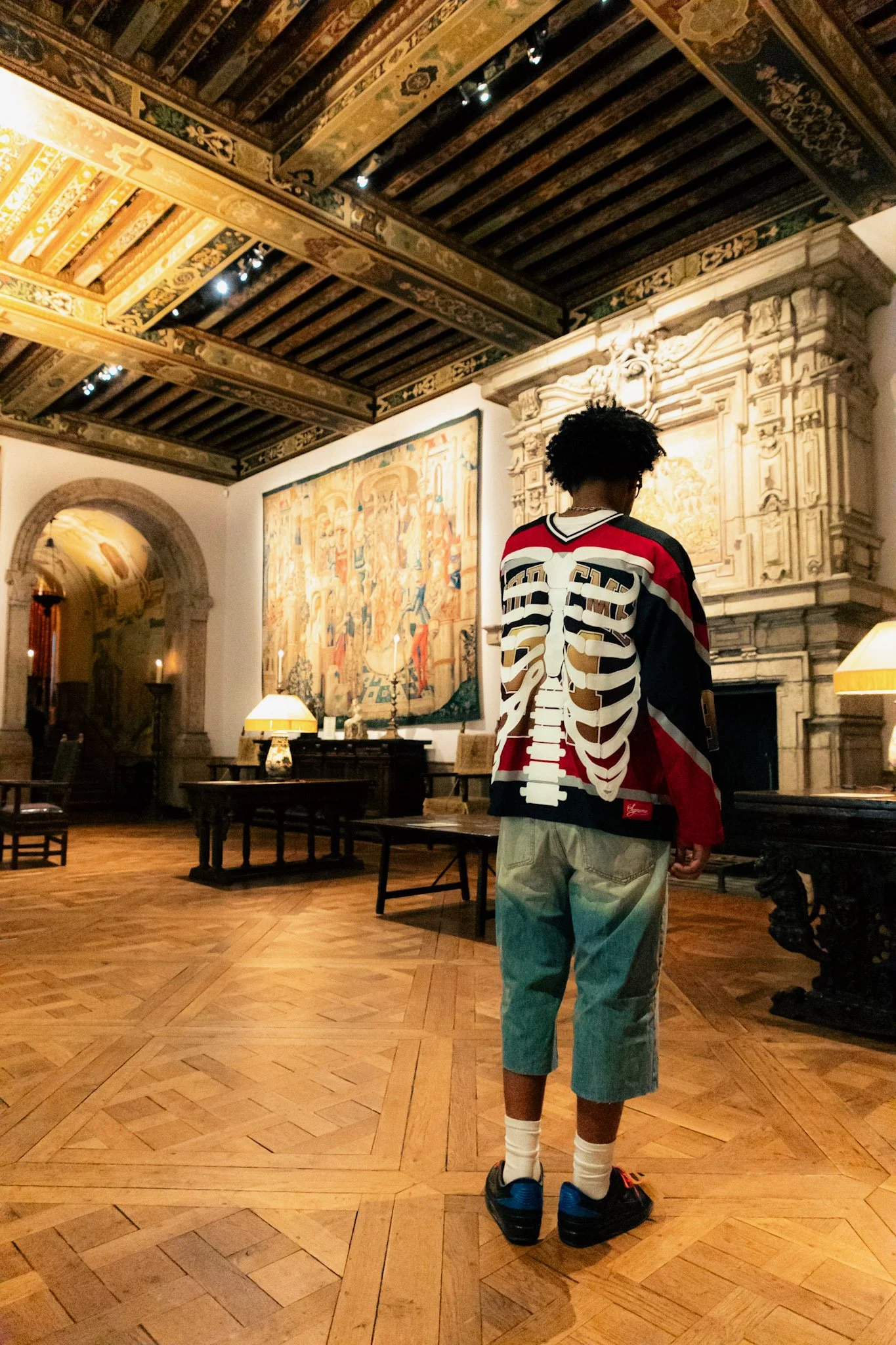A person wearing a jacket with a skeleton print on the back standing inside an ornate room with a high, decorated wooden ceiling, a large painting, and vintage furniture.