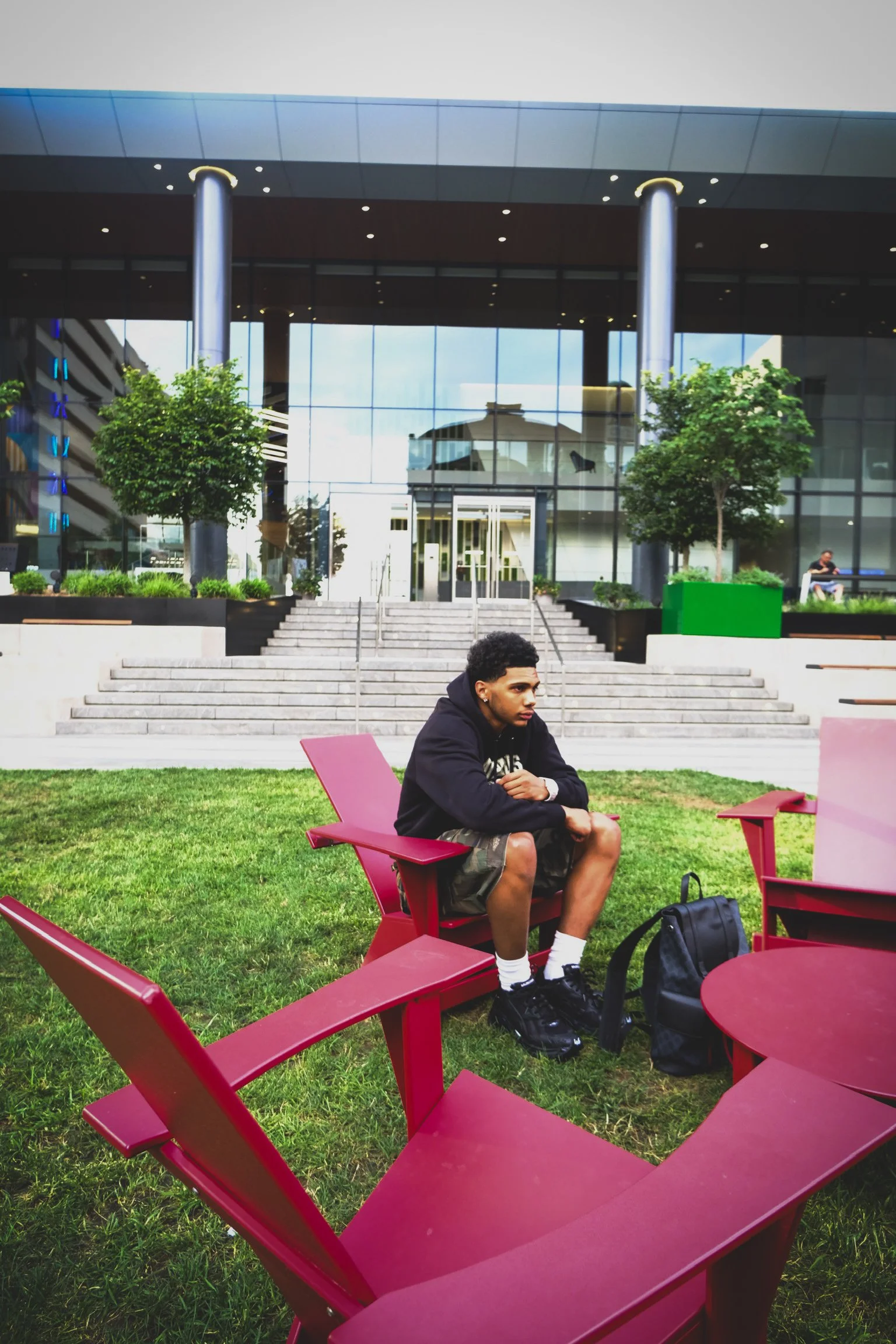 A young man sitting alone on pink outdoor chairs on a grassy area in front of a modern glass building with stairs, trees, and a person in the background.
