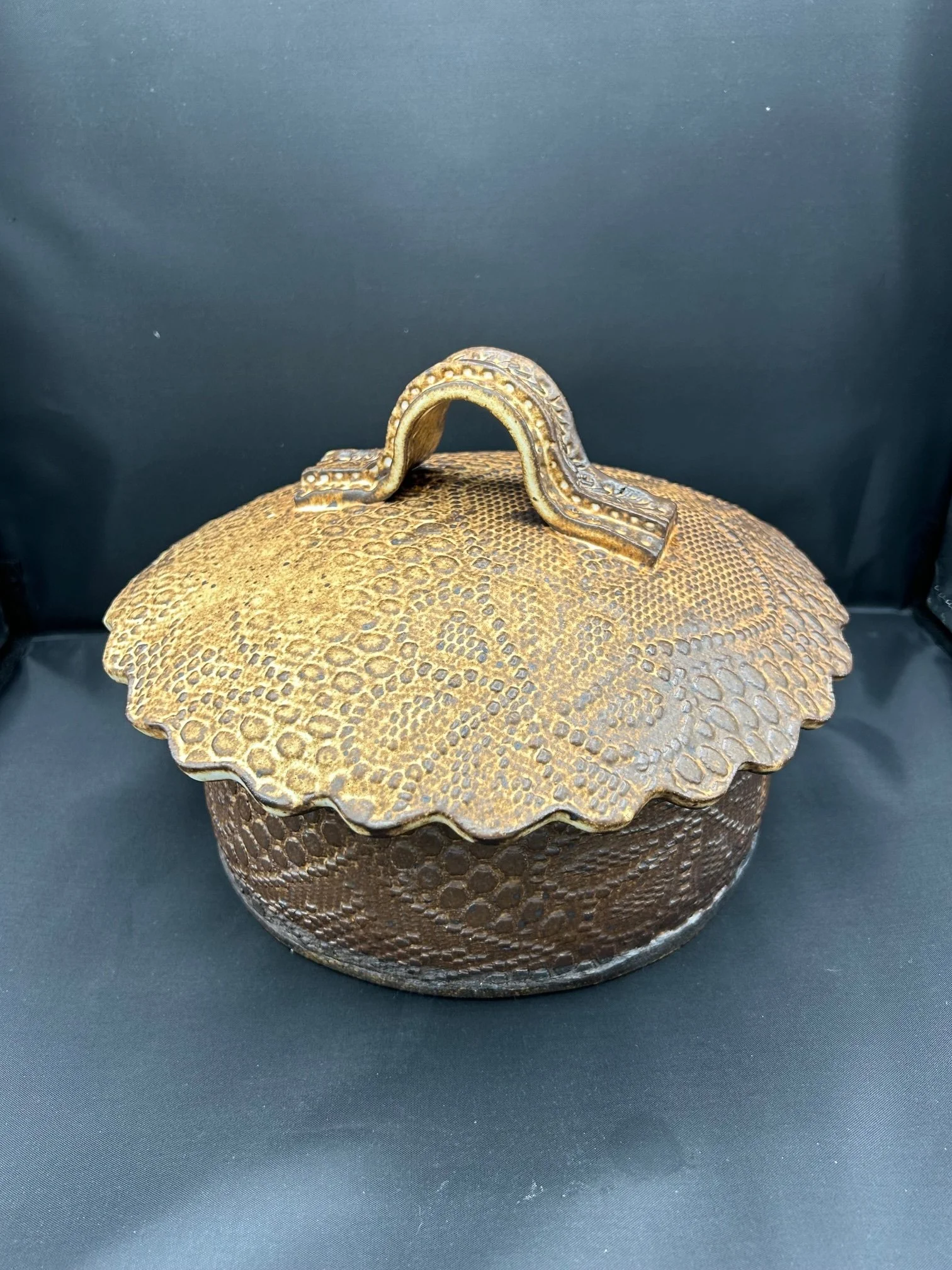 A decorative round metal container with a textured, honeycomb pattern and a scalloped edge lid, featuring a curved handle on top.