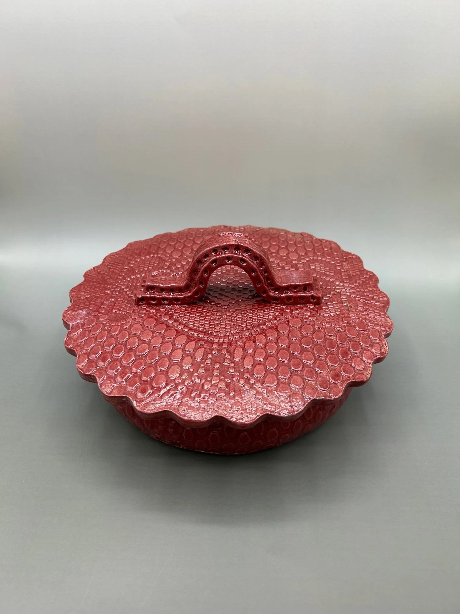 A small, round, red container with a textured, snakeskin-like pattern, featuring a handle on top, placed on a plain, light gray surface.