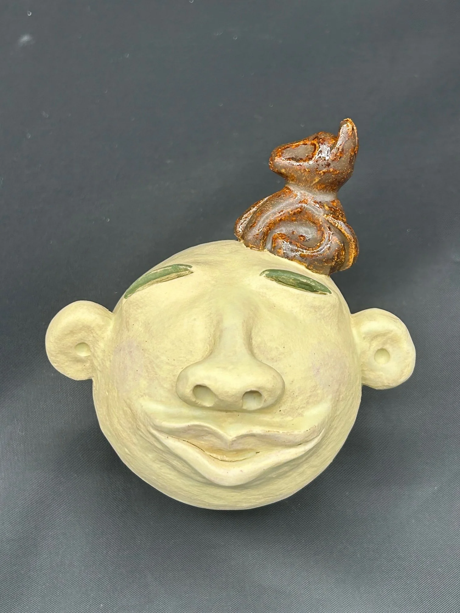 A decorative ceramic face sculpture with a smiling expression, closed eyes, and a brown swirl object on top of its head against a dark grey background.