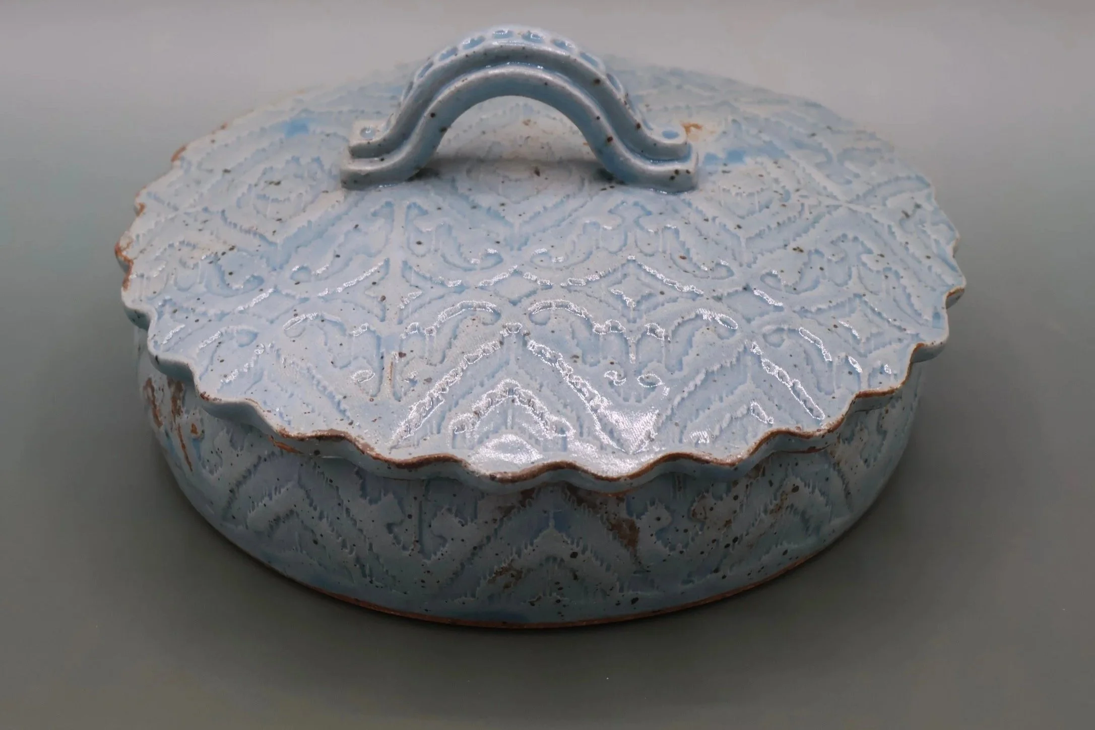 A round, vintage, decorative metal container with a white and blue floral pattern, a scalloped edge, and a curved metal handle on top.