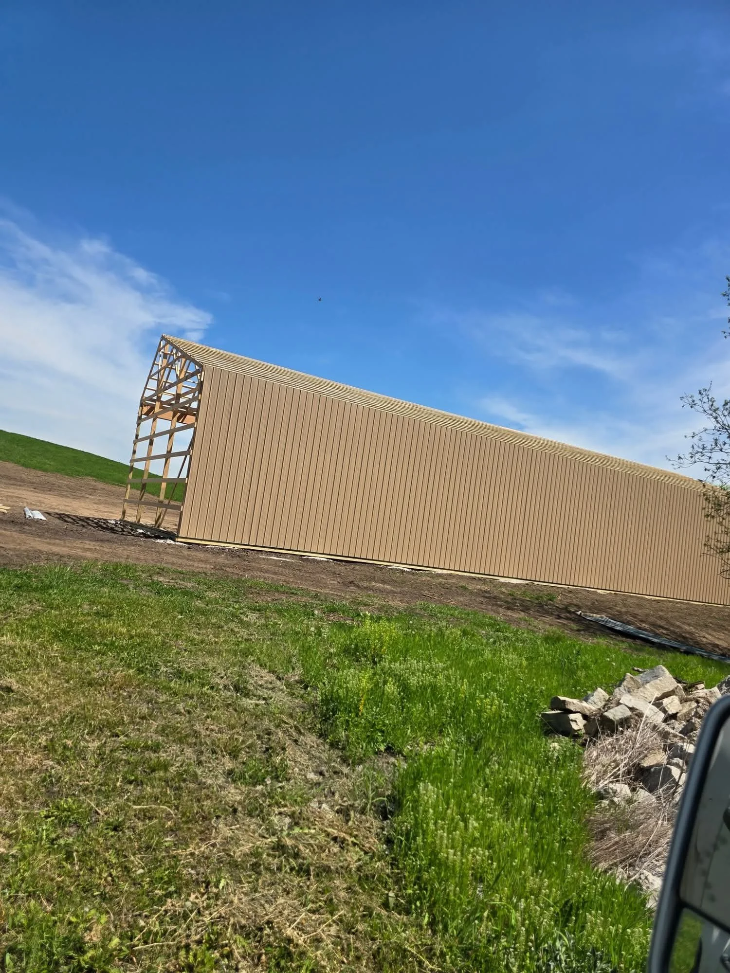 Construction site with a partially built tan-colored metal building and a grassy area with some rocks in the foreground, under a blue sky.