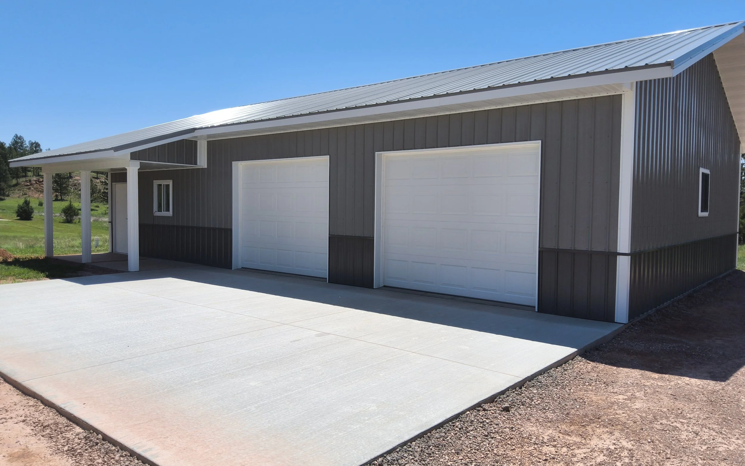 A modern detached garage with two white doors and a small window, surrounded by a concrete driveway and a grassy landscape under a clear blue sky.