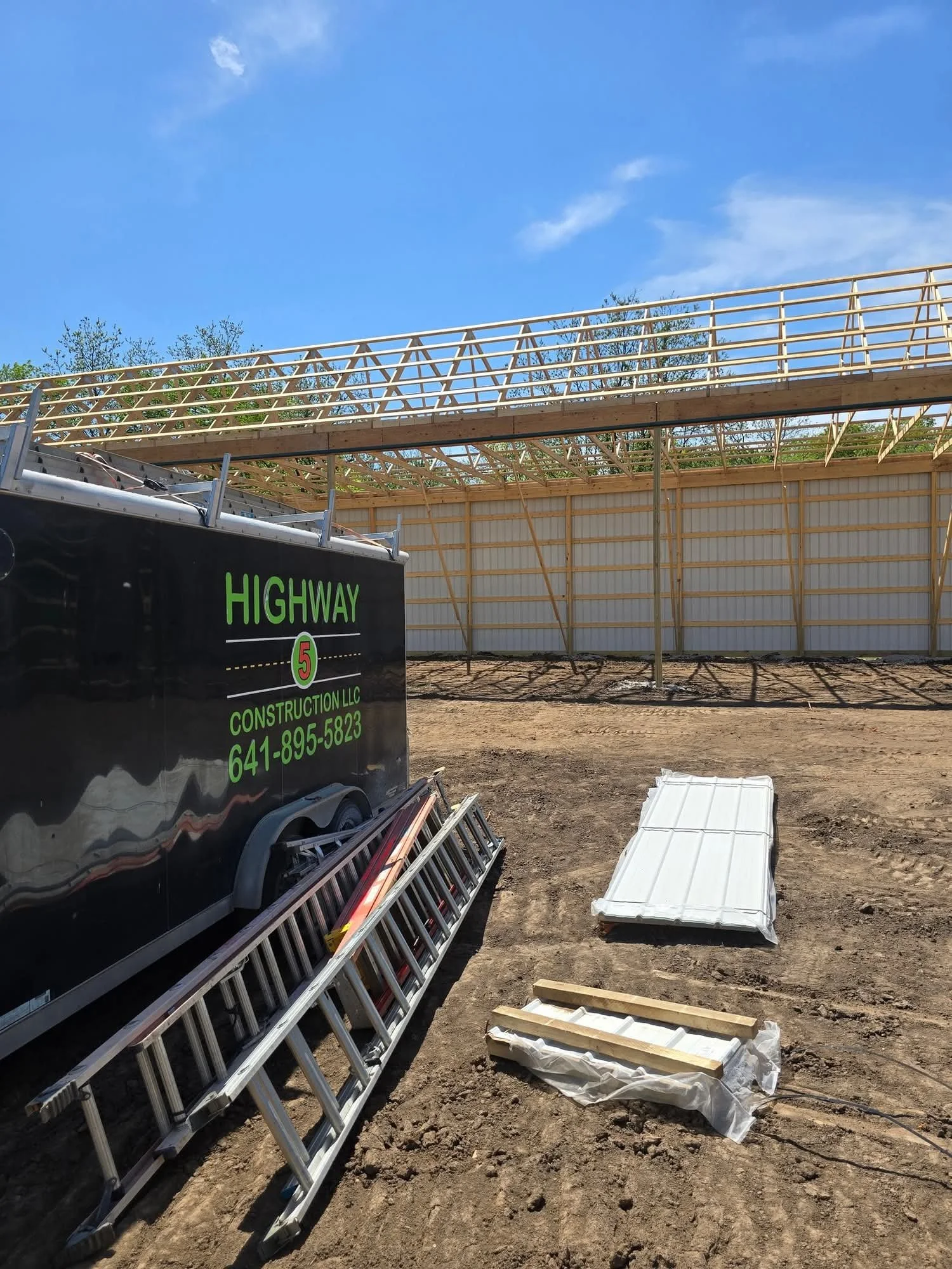 Construction site with a ladder, metal panels, and wooden framing in progress. Clear blue sky and trees in the background.