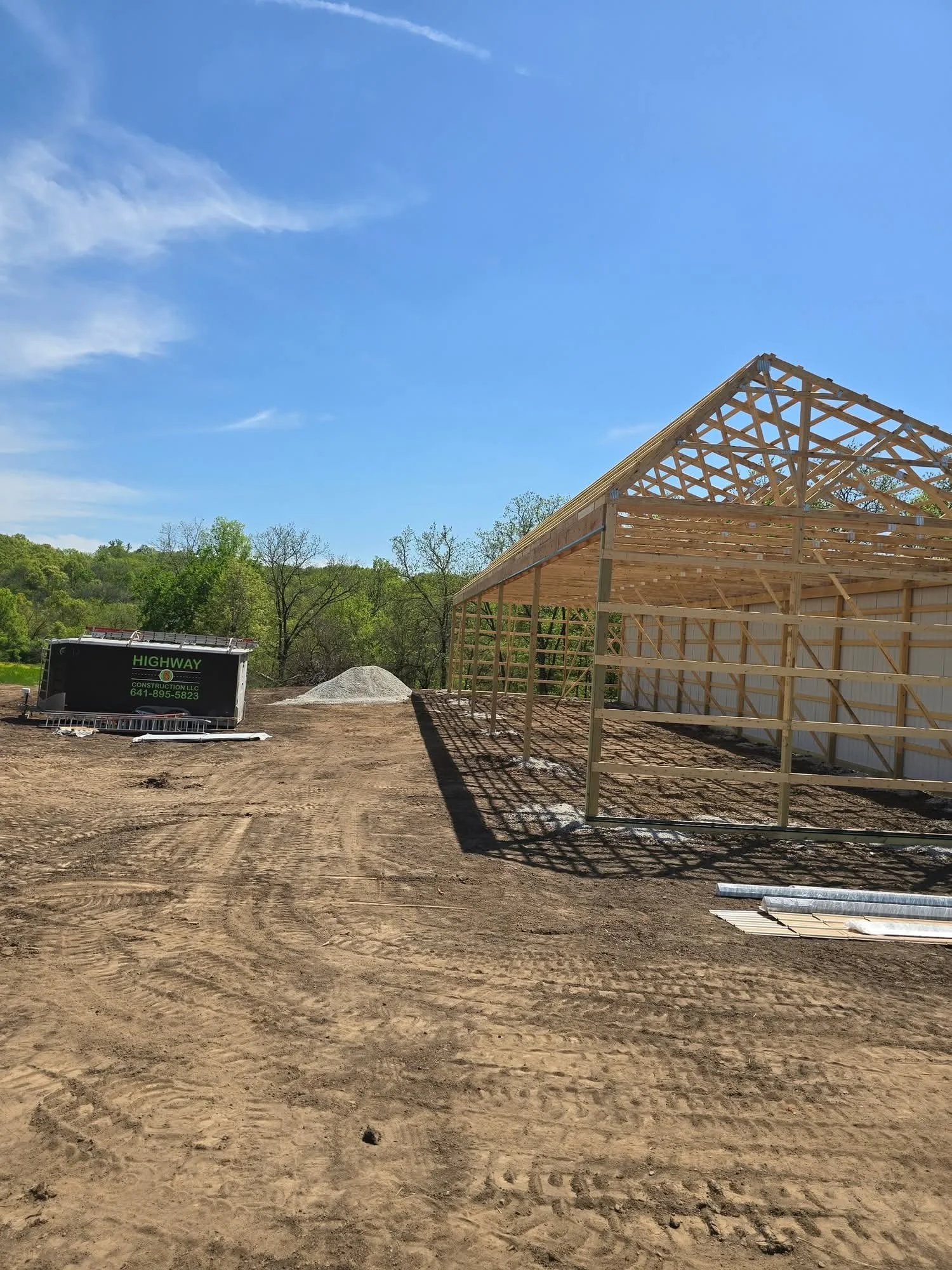 Construction site with partially built wooden structure, dirt ground, and a trailer on a sunny day with blue sky and trees in the background.