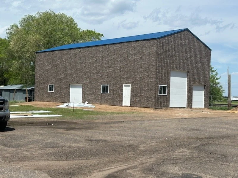 Brick building with blue metal roof, four doors, and four small windows, with some construction materials nearby, in an outdoor area with trees and cloudy sky.