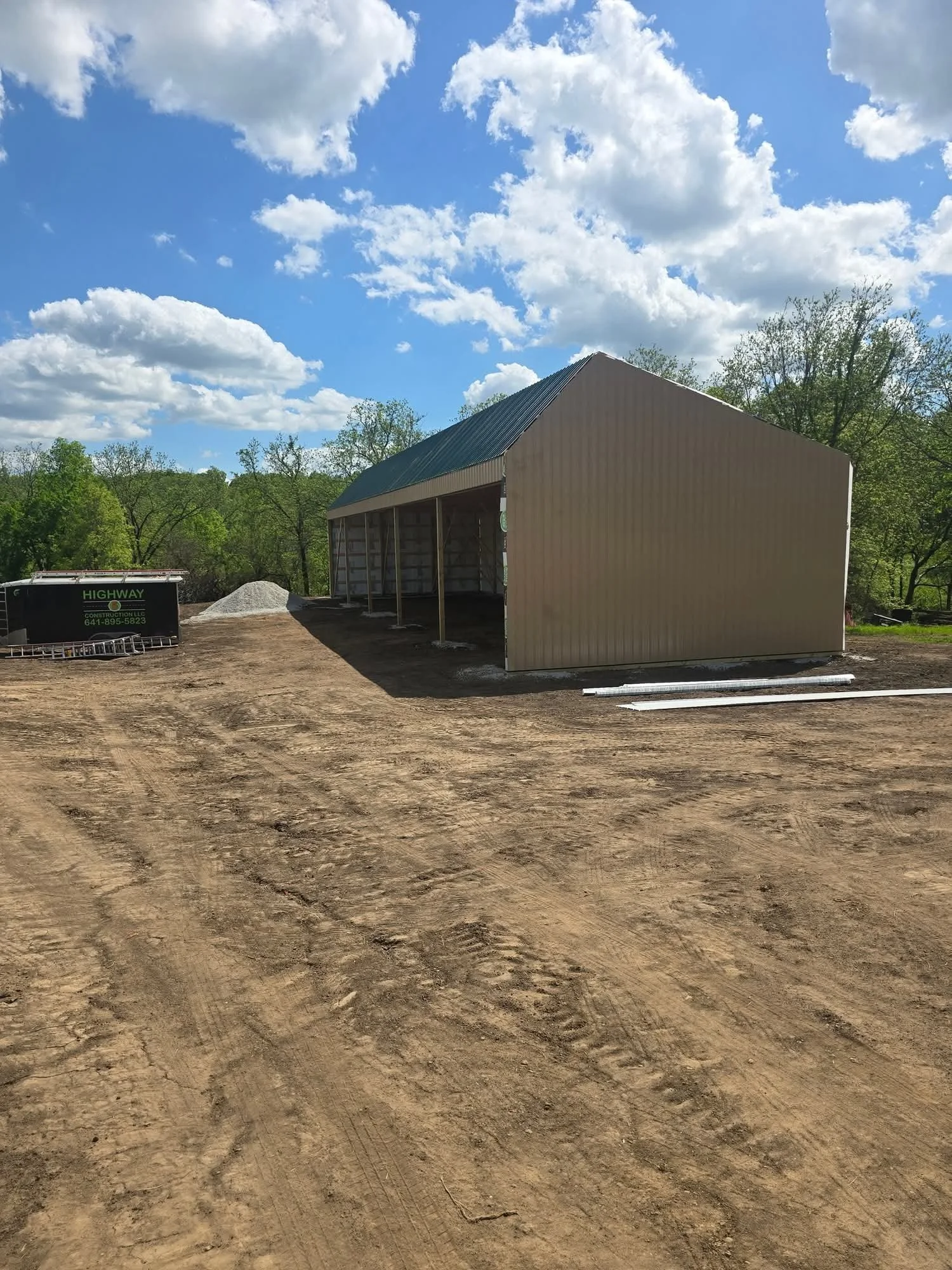 Construction site with a partially built metal building, dirt ground, a pile of gravel, and a black truck with green lettering under a partly cloudy sky.