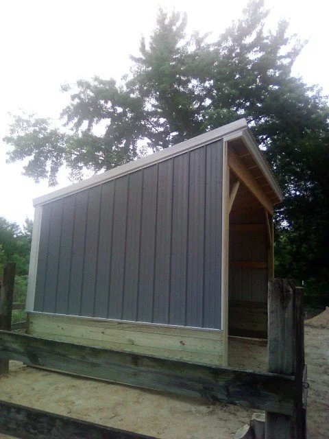 Small outdoor shed with gray siding and a sloped roof, surrounded by trees and a wooden fence in the foreground.