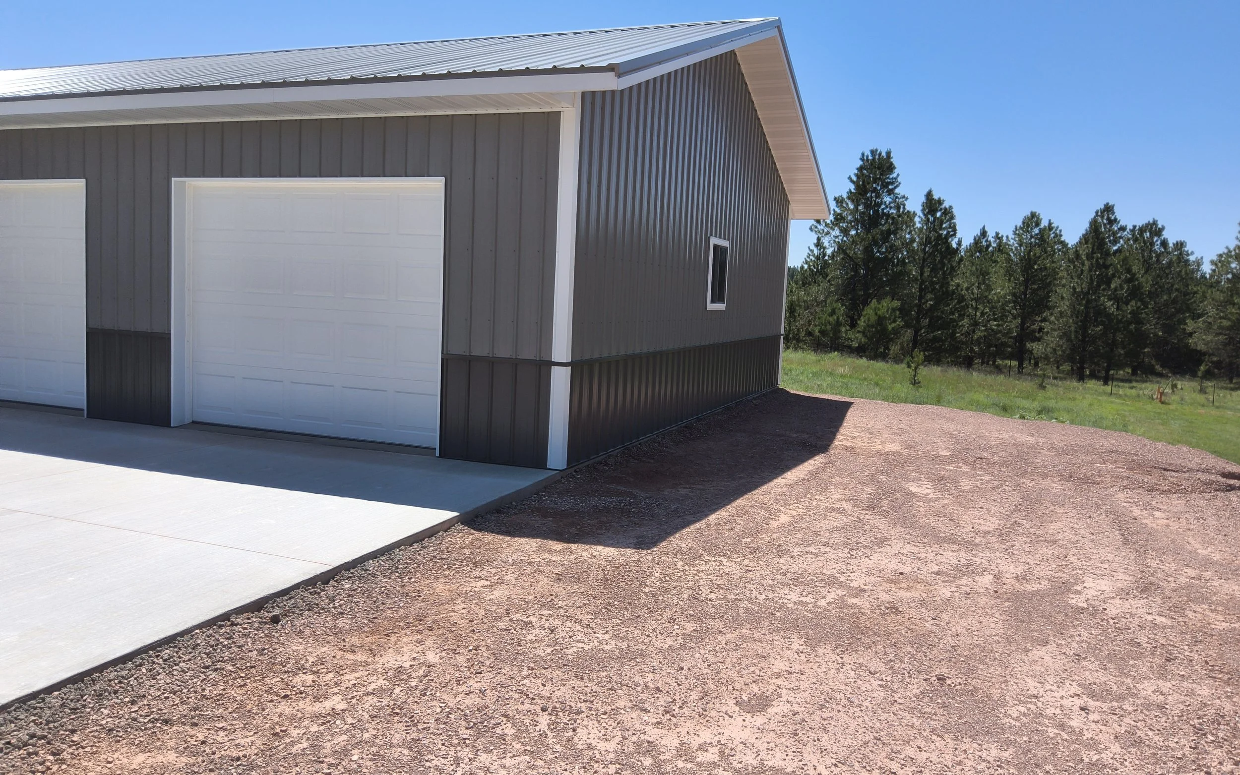 A large garage with white doors, gray metal siding, and a sloped metal roof, situated on a gravel driveway with green trees in the background under a clear blue sky.