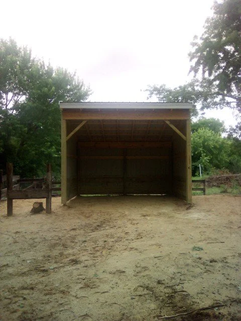 A small, open wooden shelter with a pitched roof on a dirt ground, surrounded by trees and a wooden fence.