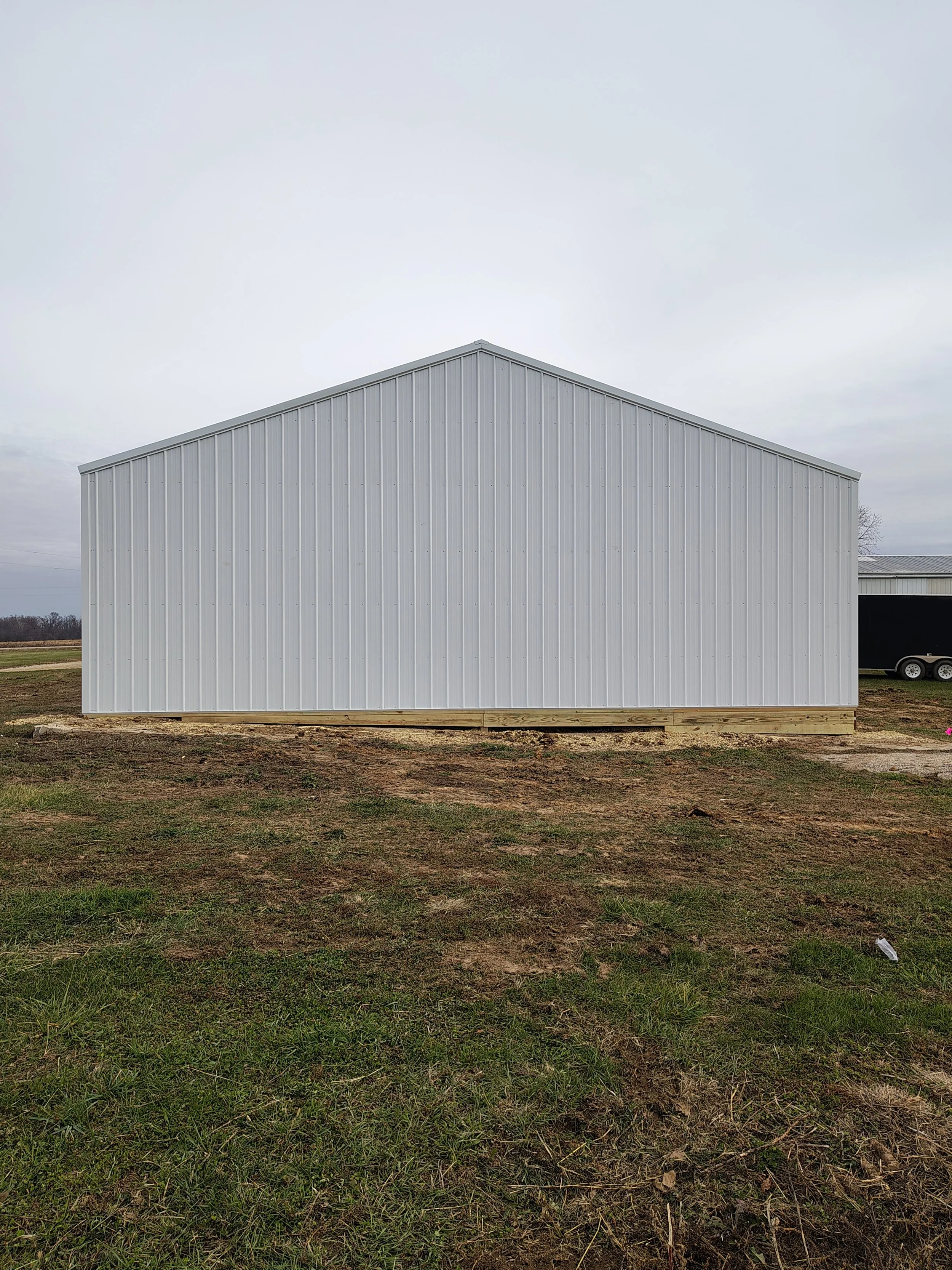 A plain white metal storage shed with vertical ridges on its exterior, located on a grassy and partially muddy area with an overcast sky.