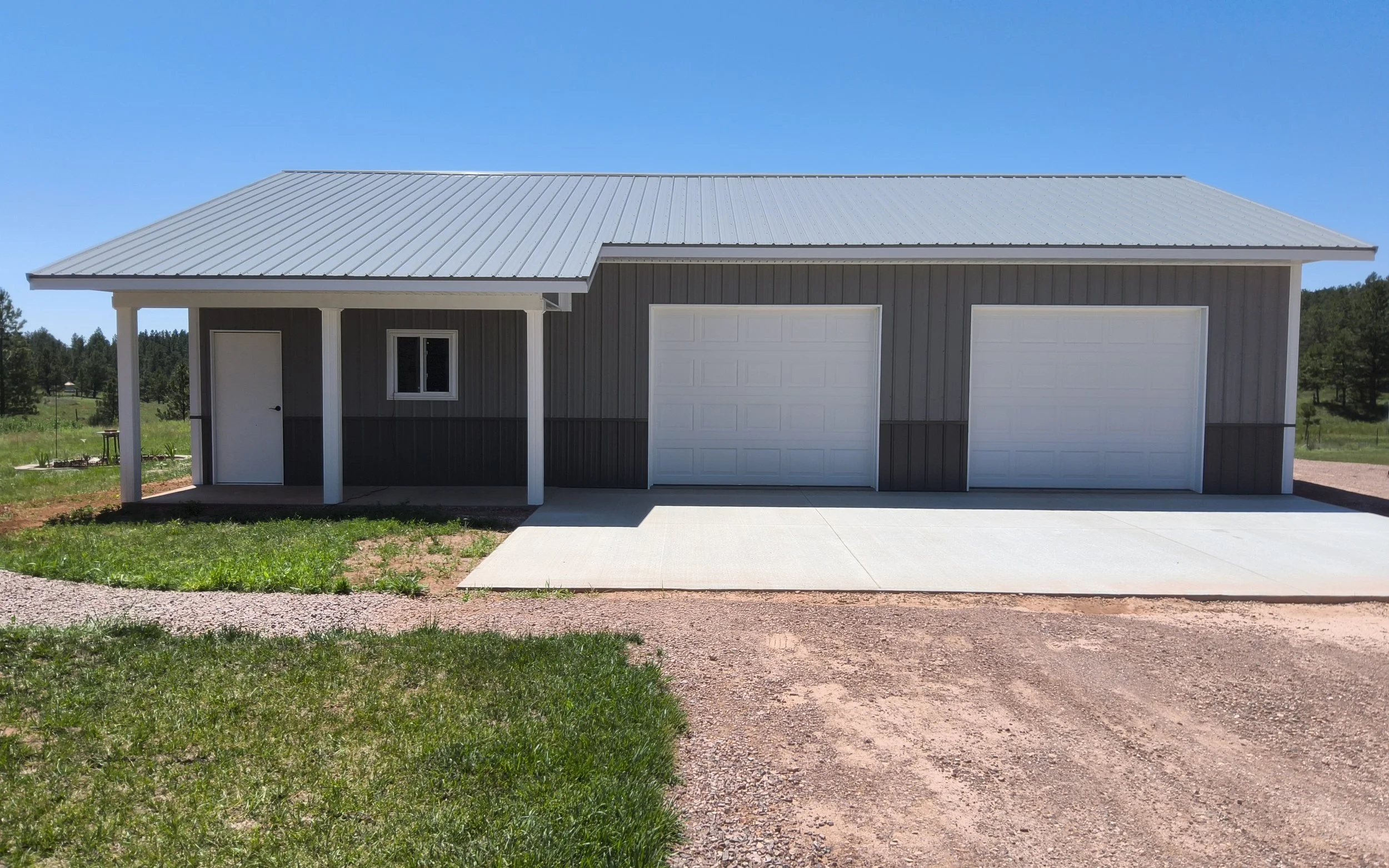 A grey metal garage building with two garage doors and a side door, situated on a concrete driveway with grass and gravel surrounding it, under a clear blue sky.
