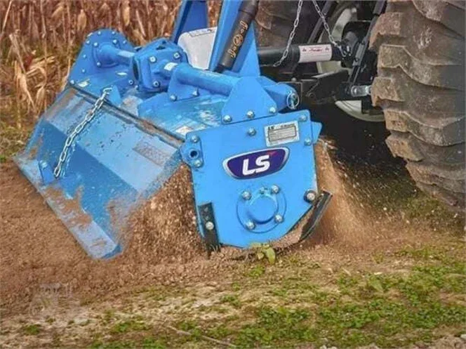 A blue LS-branded heavy-duty rototiller attached to a tractor tilling the soil.