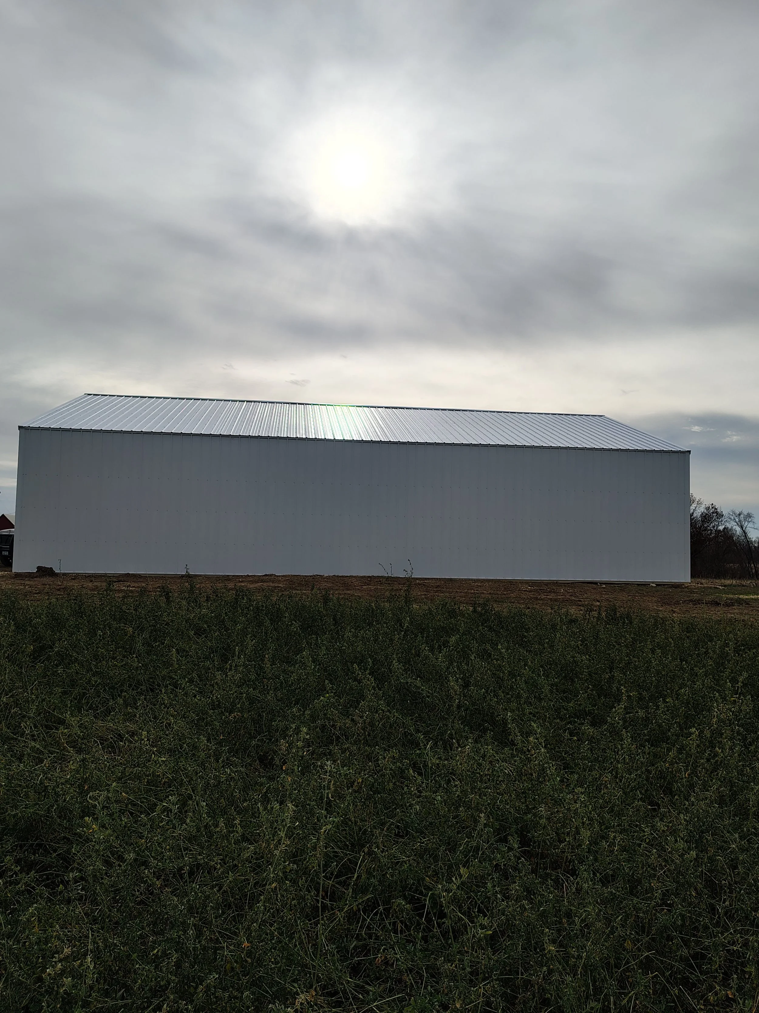 Large white metal barn with a sloped roof, situated on a patch of grass under a cloudy sky with the sun partially visible.