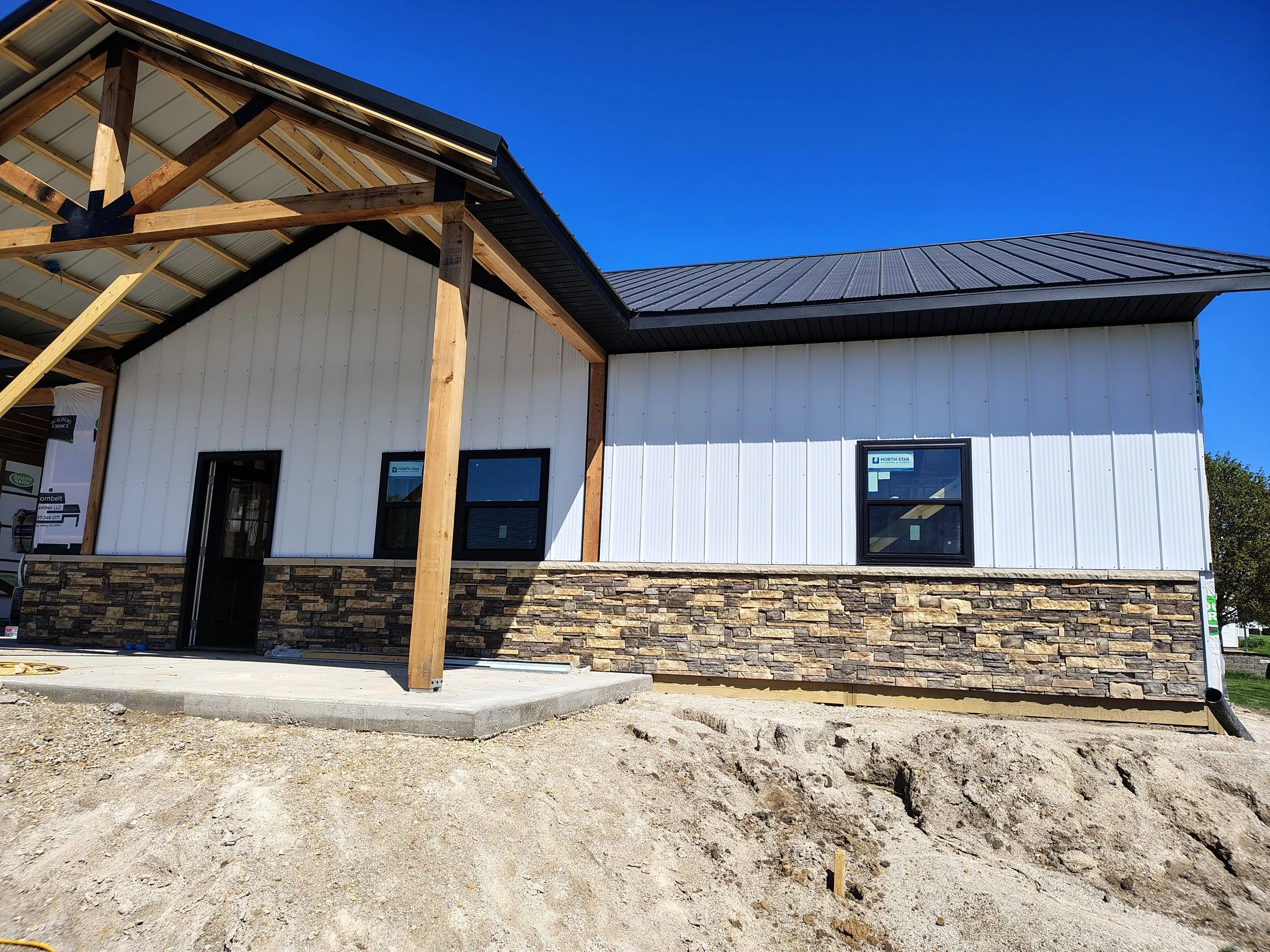A house under construction with a mix of white siding and stone exterior, black-framed windows, a sloped black metal roof, and a front porch with a concrete slab, surrounded by dirt and construction materials.