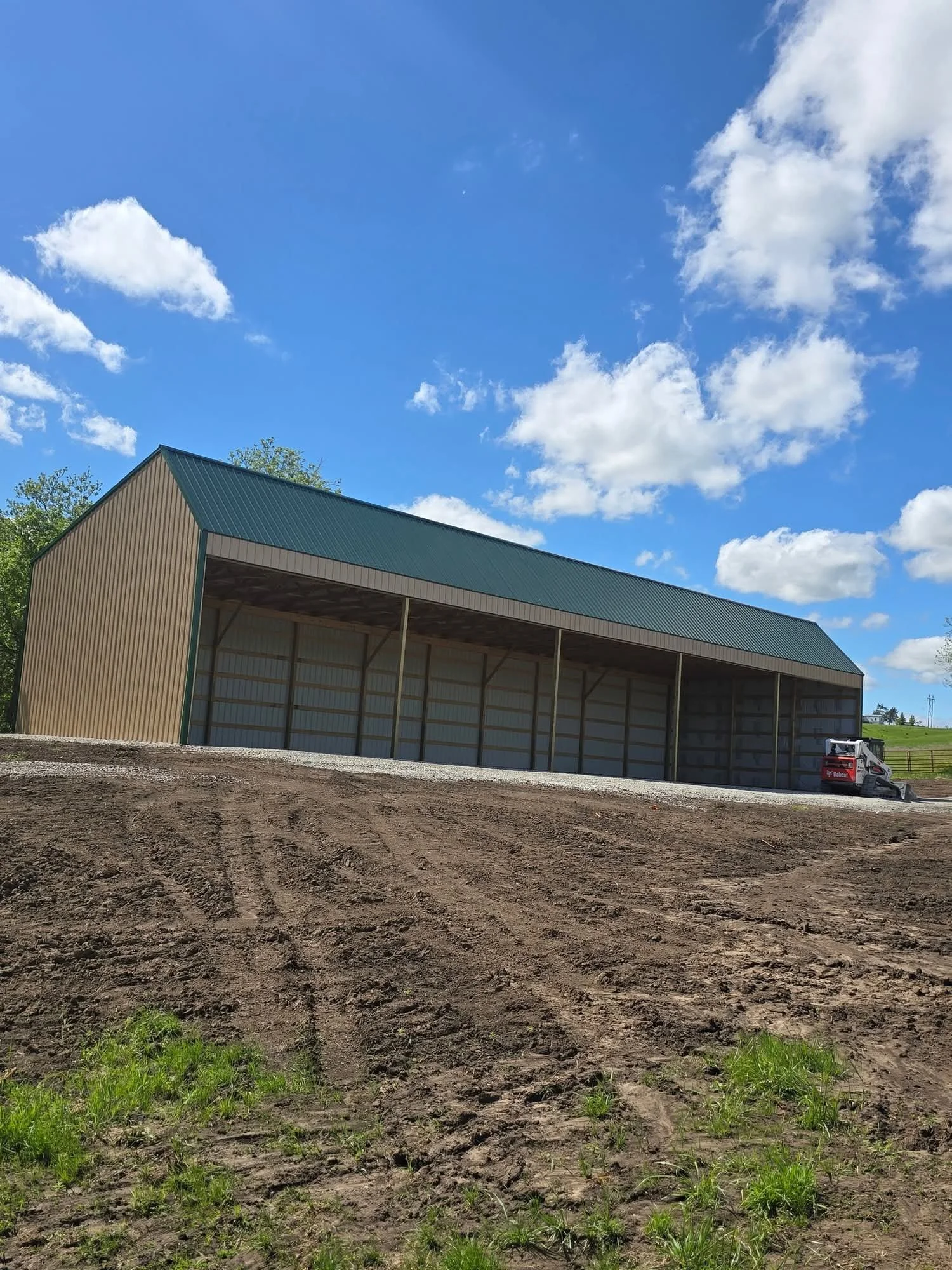 Large metal barn under construction on a dirt lot with a blue sky and white clouds overhead. A small black and red piece of equipment is visible to the right.