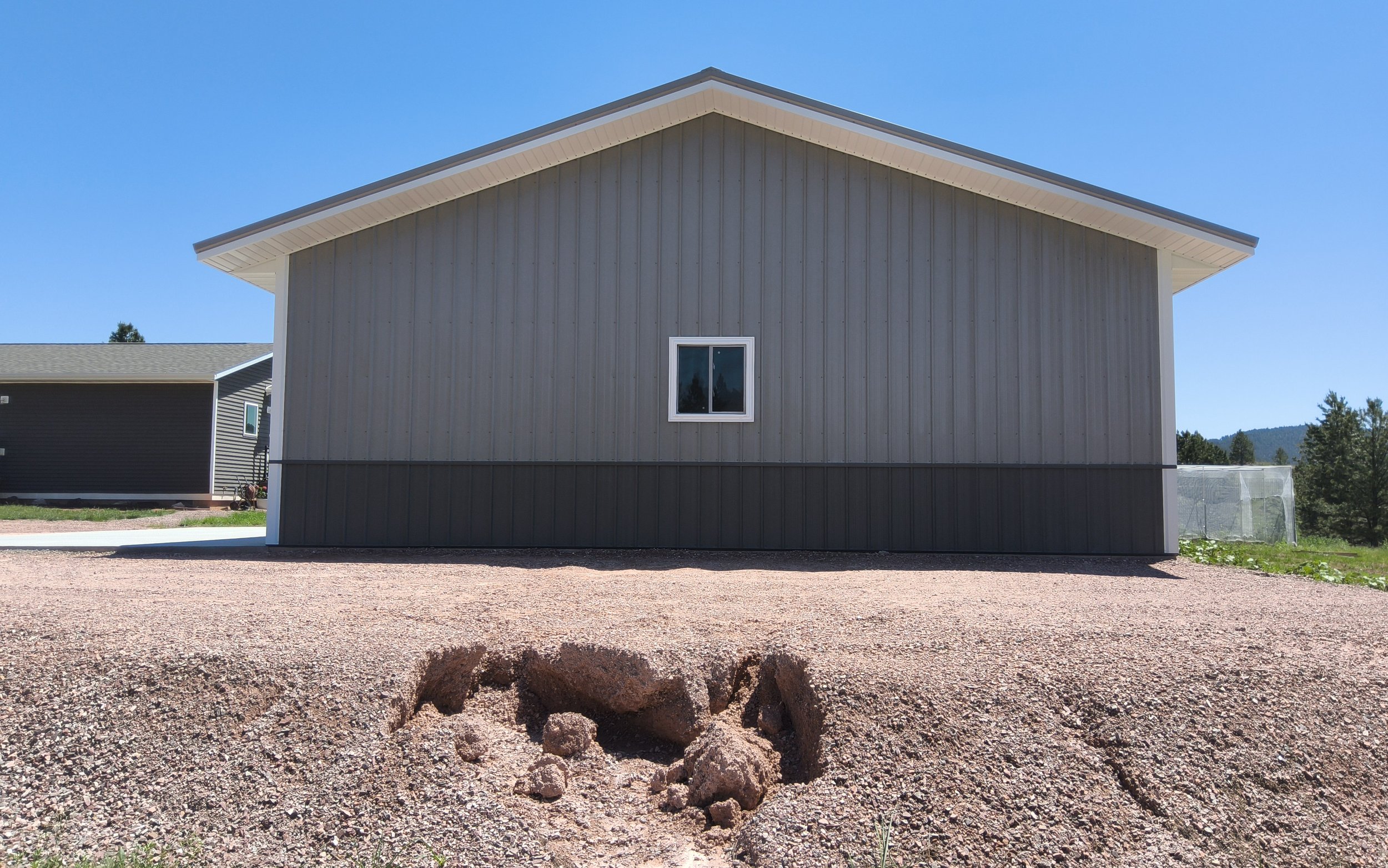 A house with gray siding and a white frame window on a gravel lot under a clear blue sky, with a large hole and rocks in the foreground.