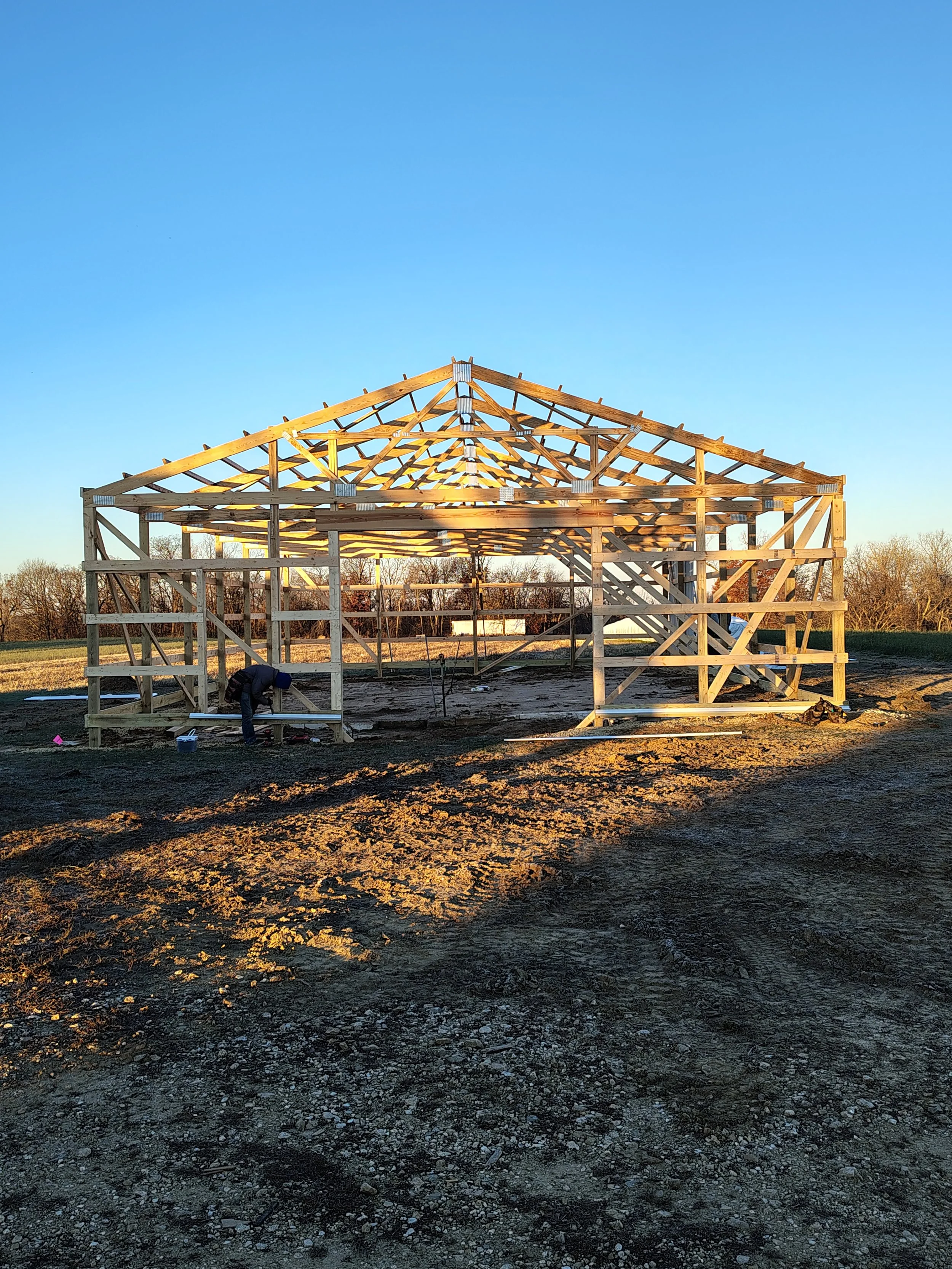 Wooden house frame under construction on dirt ground with a worker working nearby, clear blue sky in the background.
