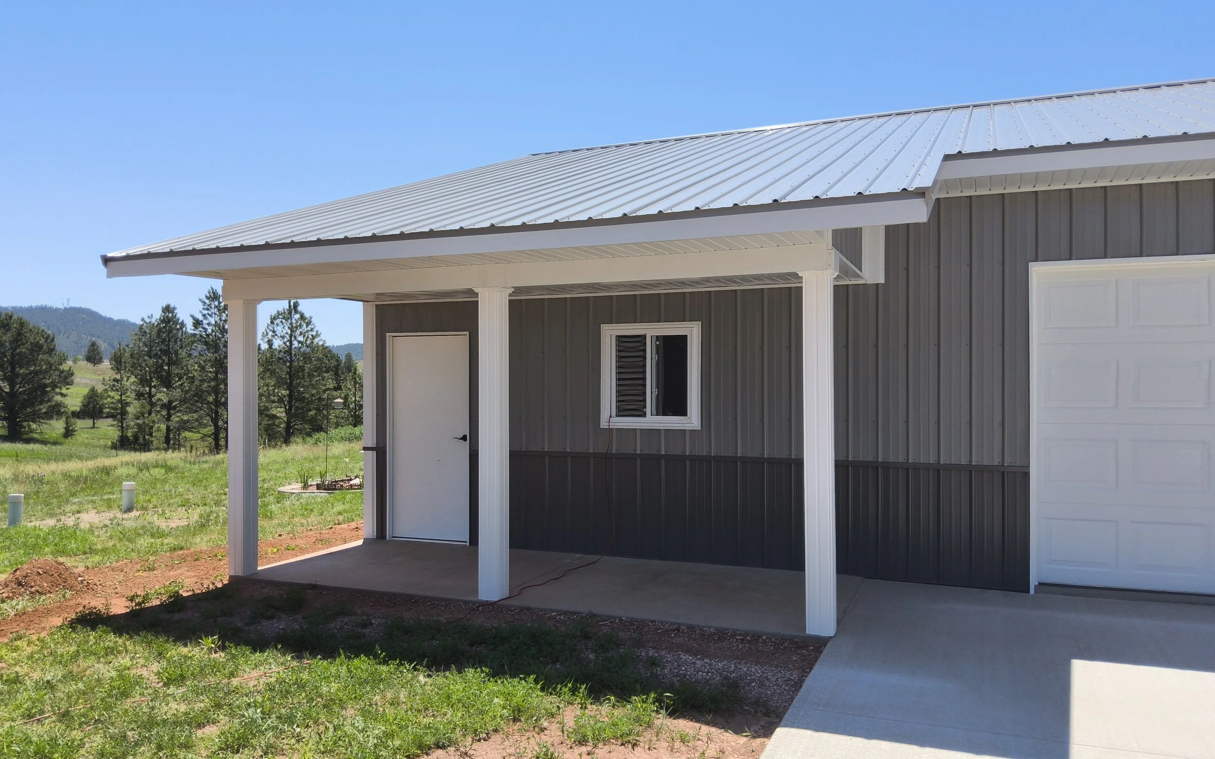 Newly constructed building with a metal roof, small porch, gray siding, white trim, a white door, a small window, and a white garage door, set against a rural landscape with green grass, trees, and distant hills under a clear blue sky.