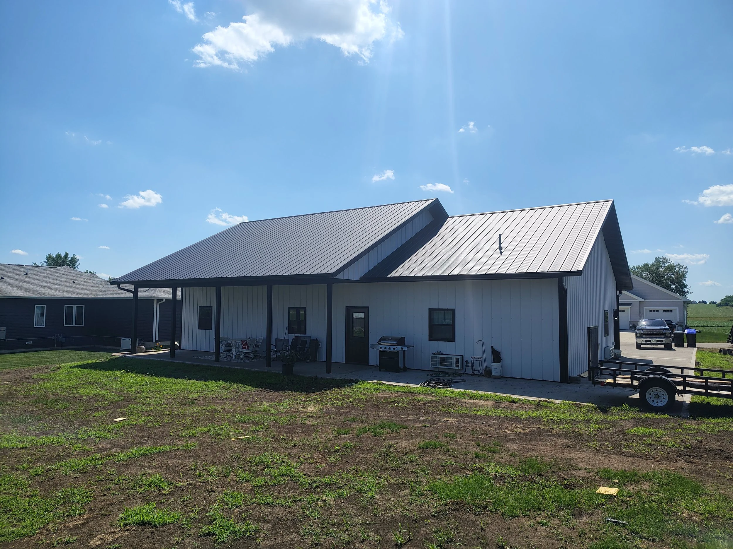 A modern metal house with a porch, outdoor furniture, barbecue grill, and a driveway with vehicles under a bright blue sky.