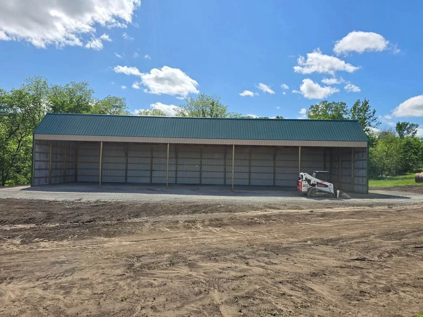 Construction site with a large metal building under construction, a mini skid steer loader parked outside, and a dirt pathway in the foreground with green trees and a blue sky with scattered clouds in the background.