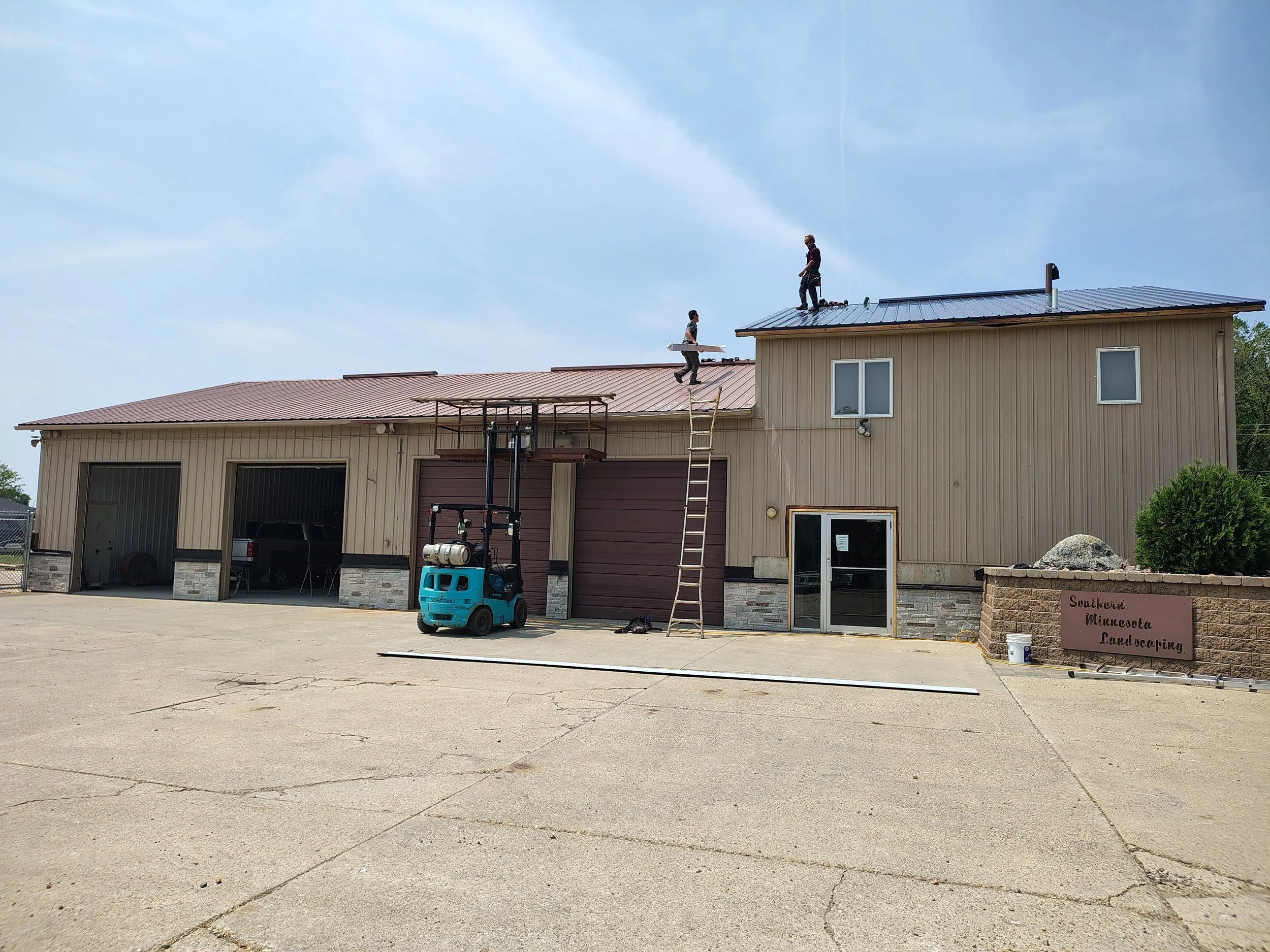 Three workers installing a metal roof on a beige building with a sign that reads 'Southwest Minnesota Landscaping' in Minnesota. The workers are on the roof, one on top and two on the edge, with ladders and a blue lift.
