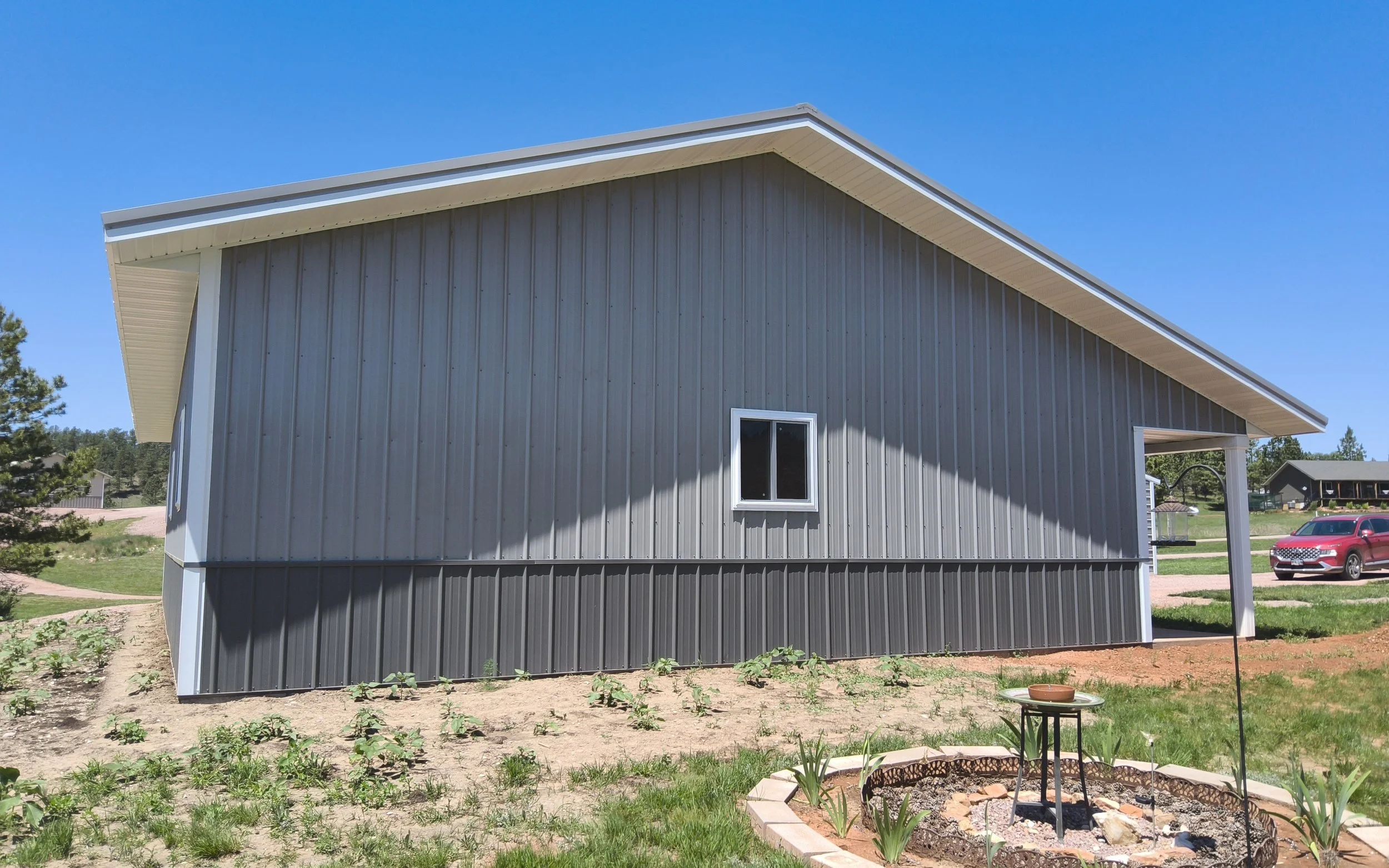 Side view of a gray metal house with a sloped roof and a small window, situated on a lawn with young plants and a circular flower bed in the foreground, under a clear blue sky.