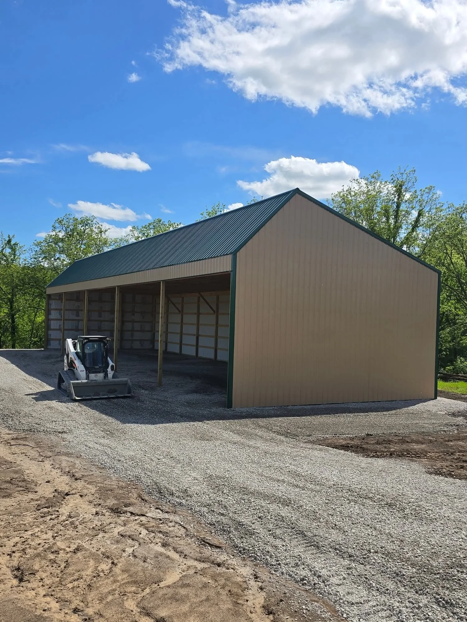 A large, metal, open-sided storage shed with a green roof, situated on a gravel area with a small Bobcat loader parked inside, surrounded by trees under a partly cloudy sky.