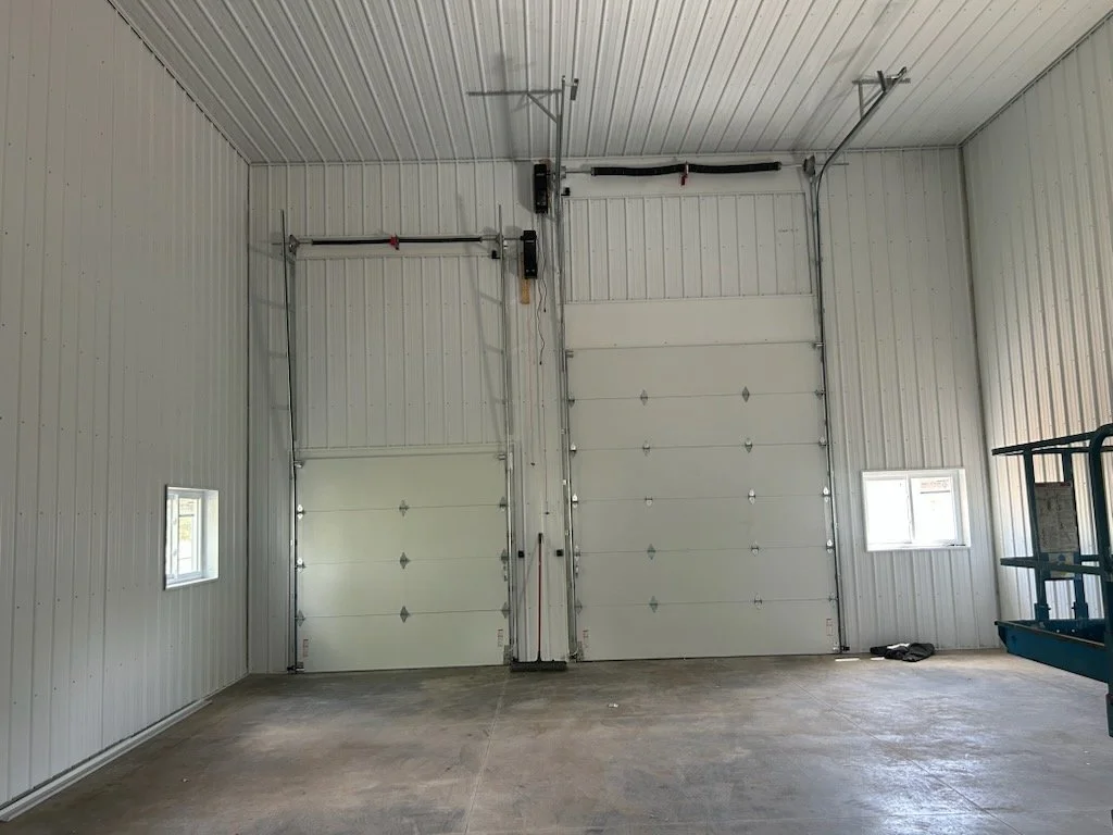 Interior of a large metal garage or warehouse with two closed white roll-up doors, two small windows, and a lift on the right side.
