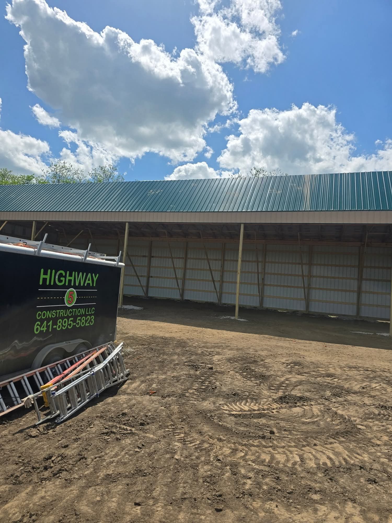 Construction site with dirt ground, a black trailer with green lettering, and a partially built metal structure with a green roof against a blue sky with clouds.