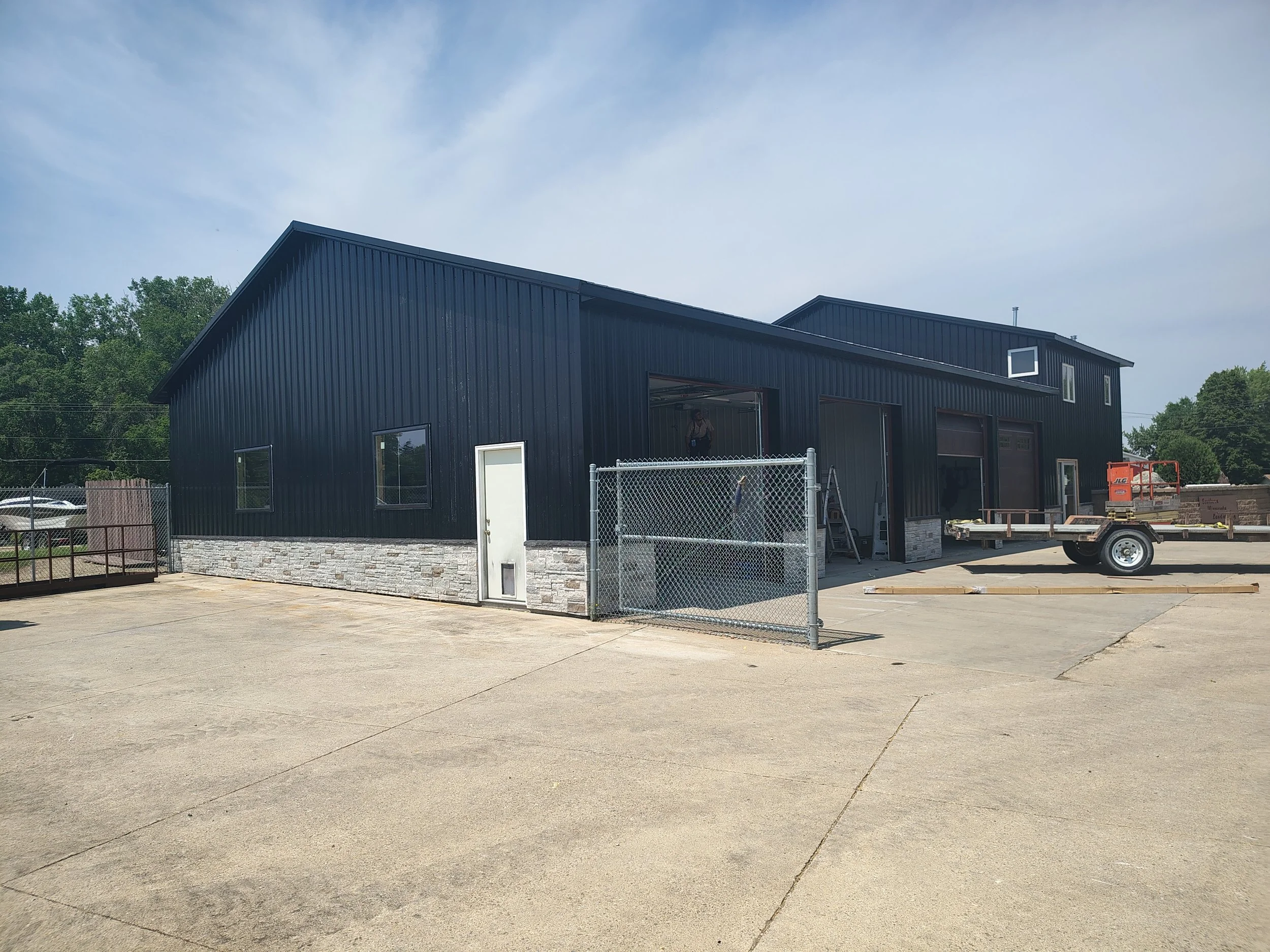 A black metal building under construction with stone accents at the base, open garage doors, a chain-link fence gate in front, and construction equipment on the concrete lot.