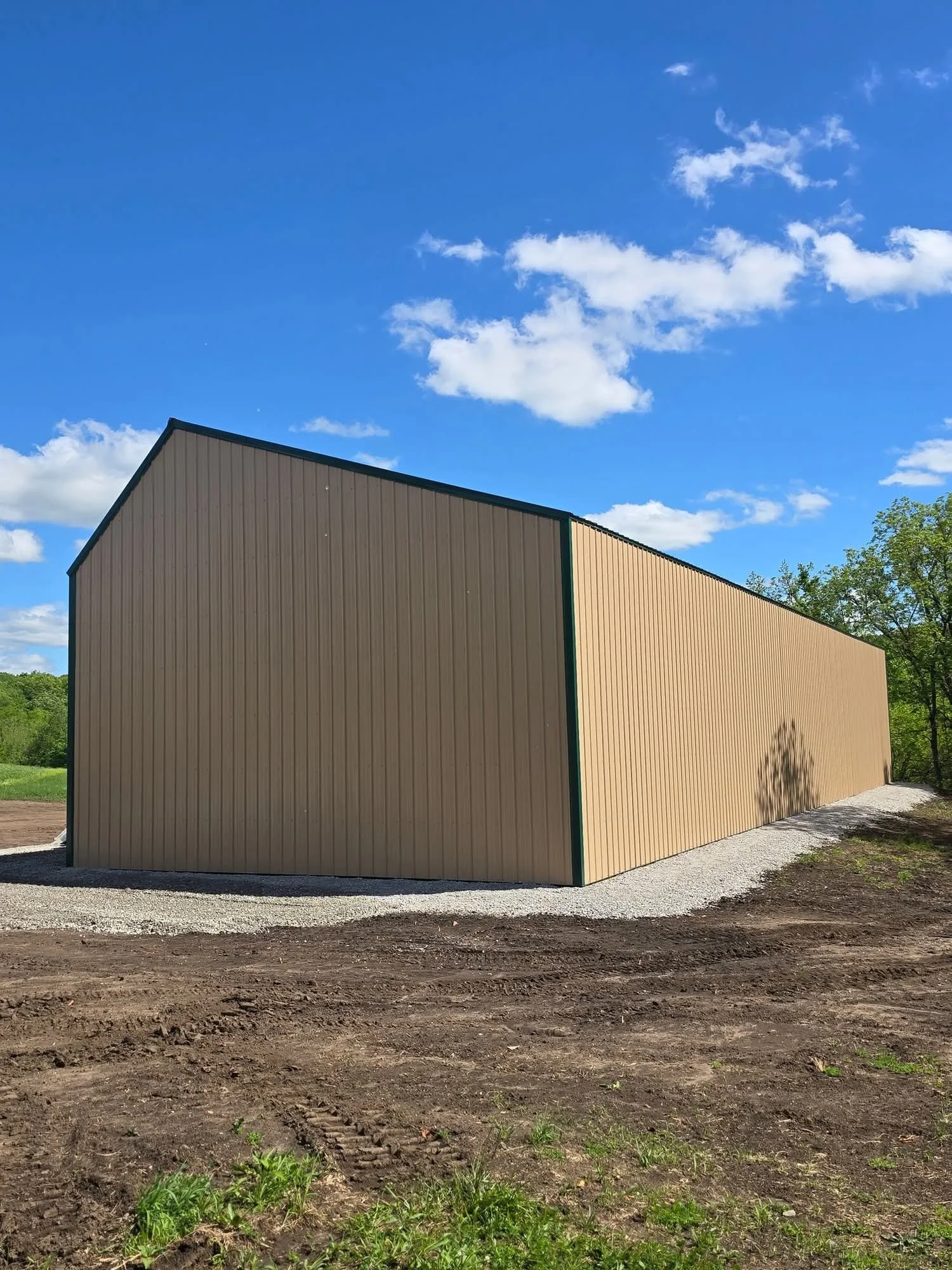 A beige metal building with a gable roof situated on a dirt ground with some green grass, under a blue sky with scattered white clouds, and in a natural, rural environment.