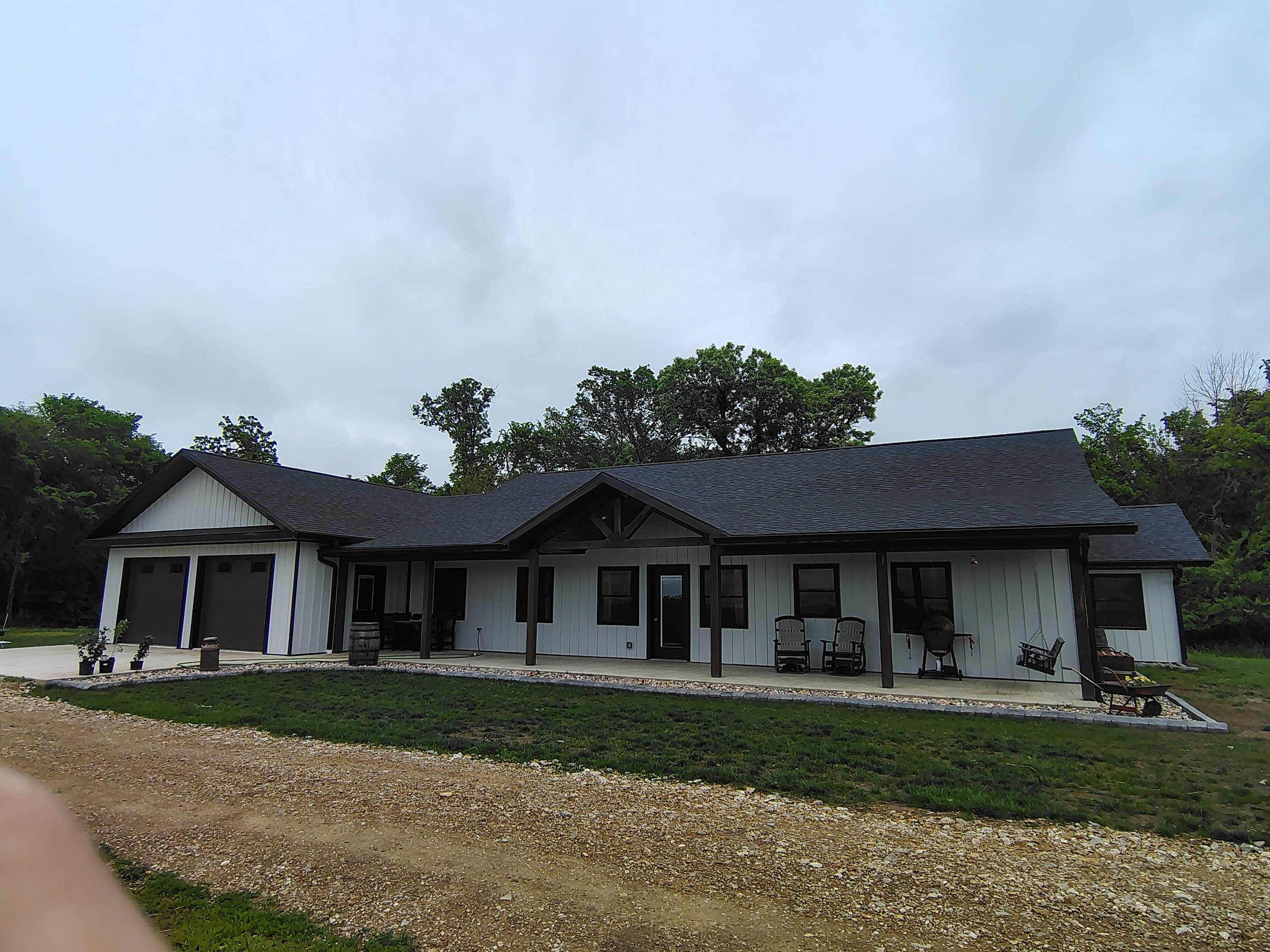 Modern farmhouse-style house with white exterior, black roof, and black trim, featuring attached garage, covered porch, outdoor seating, and gravel driveway, surrounded by trees under a cloudy sky.