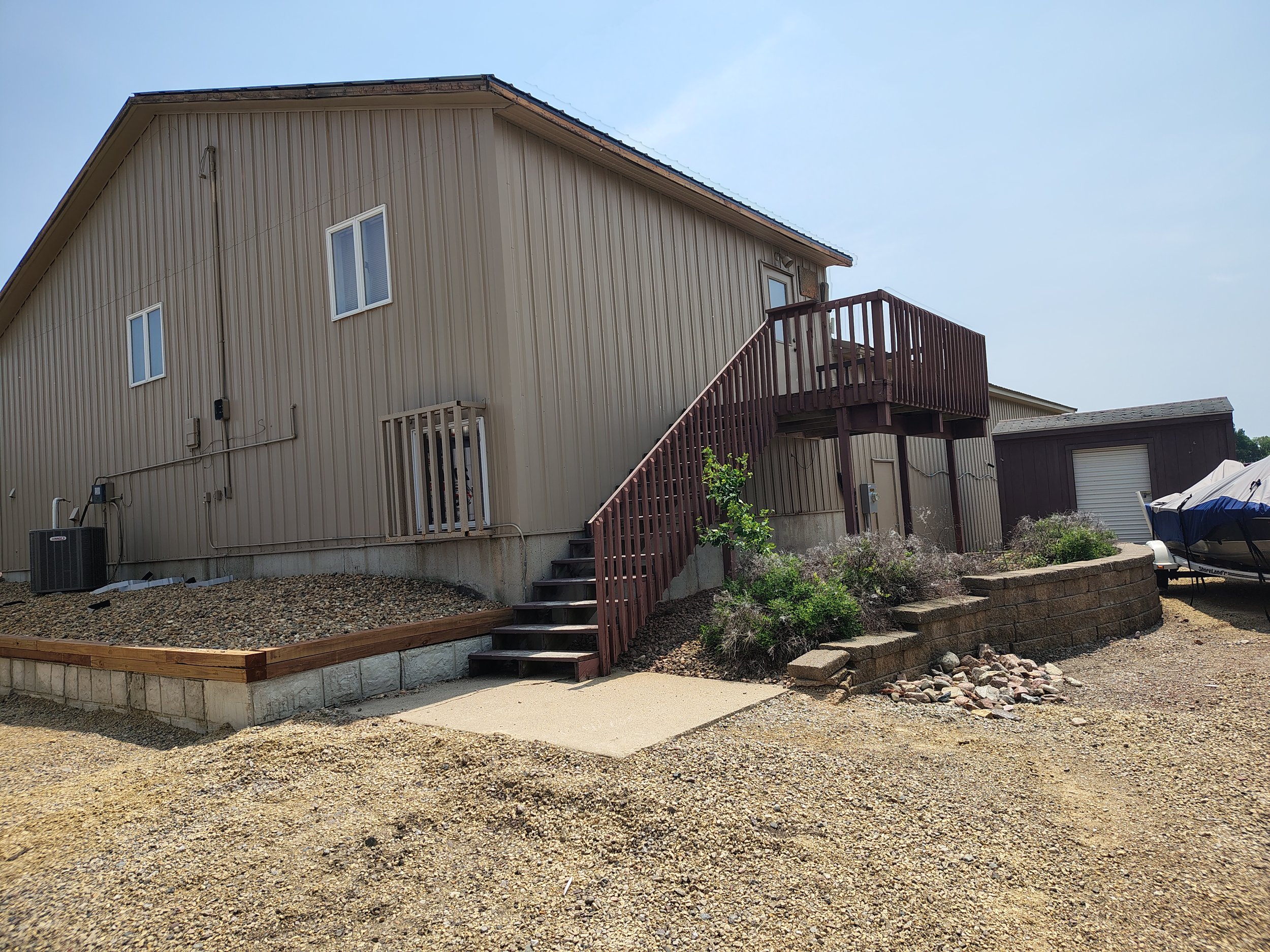 The back of a beige house with a wooden staircase leading to a deck, surrounded by gravel and landscaping with some plants, and a boat on a trailer to the right.