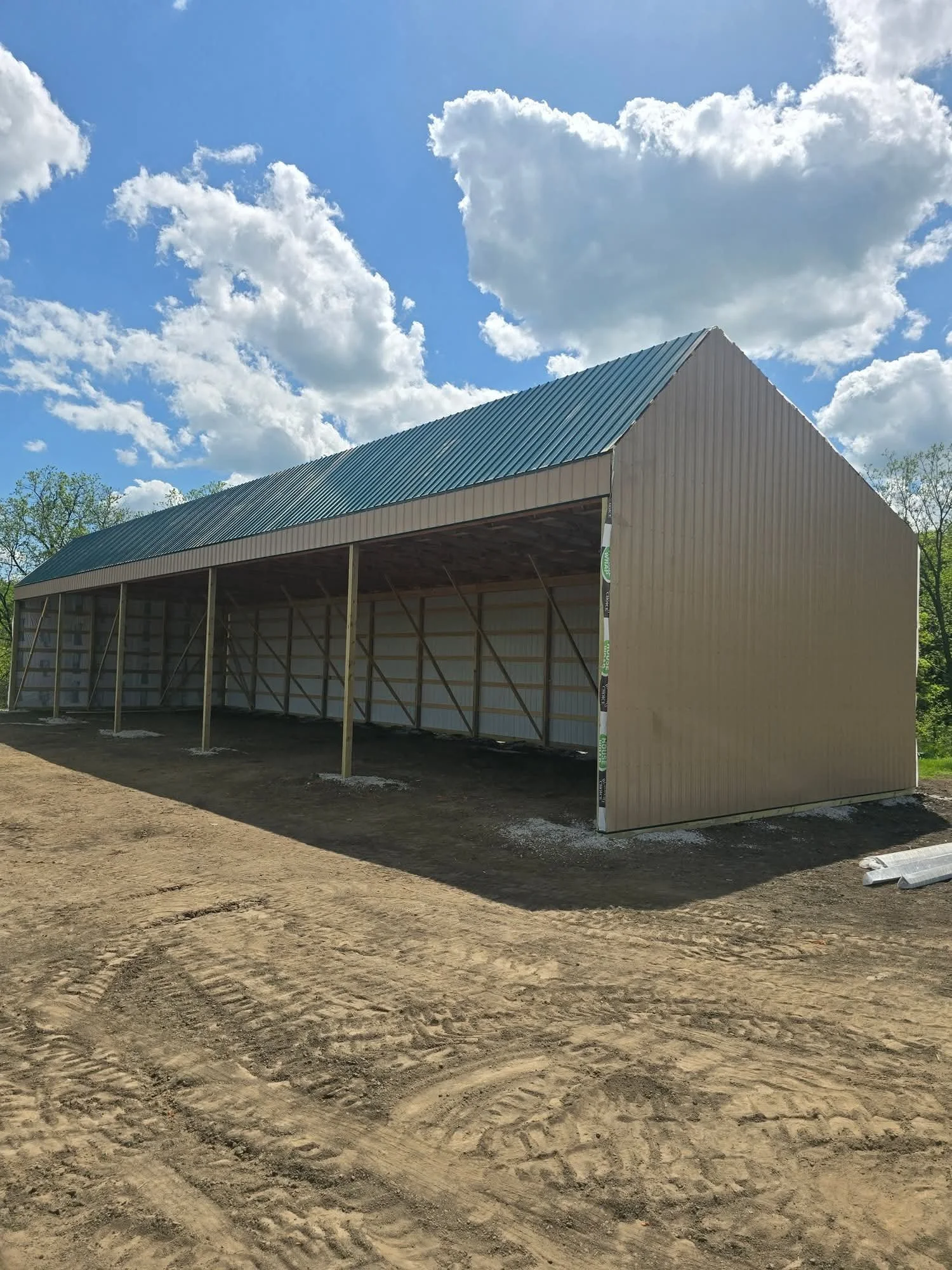 A partially built metal barn with a blue-green roof on a dirt lot under a blue sky with white clouds.