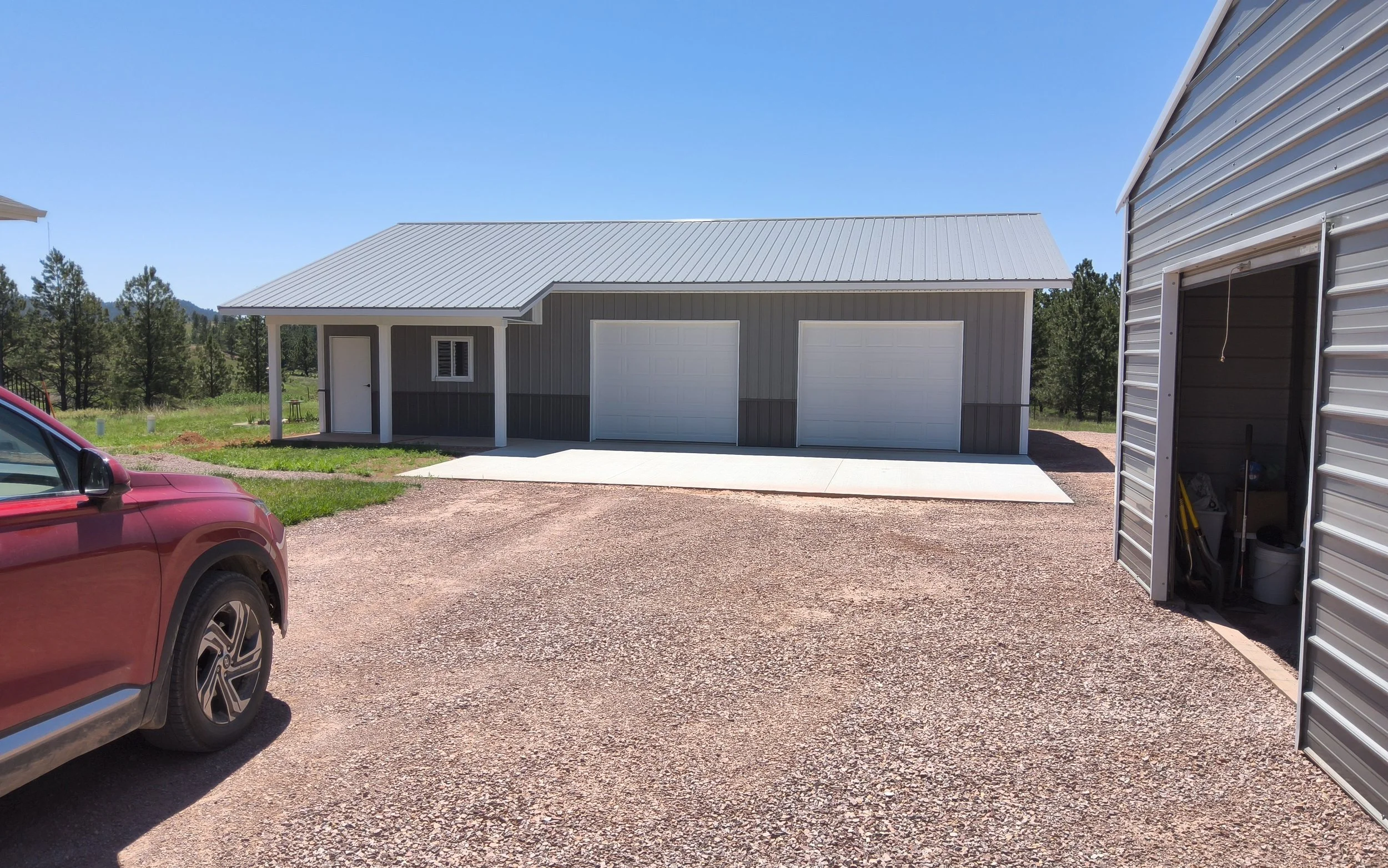 A gravel driveway in front of a gray house with a metal roof and two garage doors, surrounded by trees under a clear blue sky.