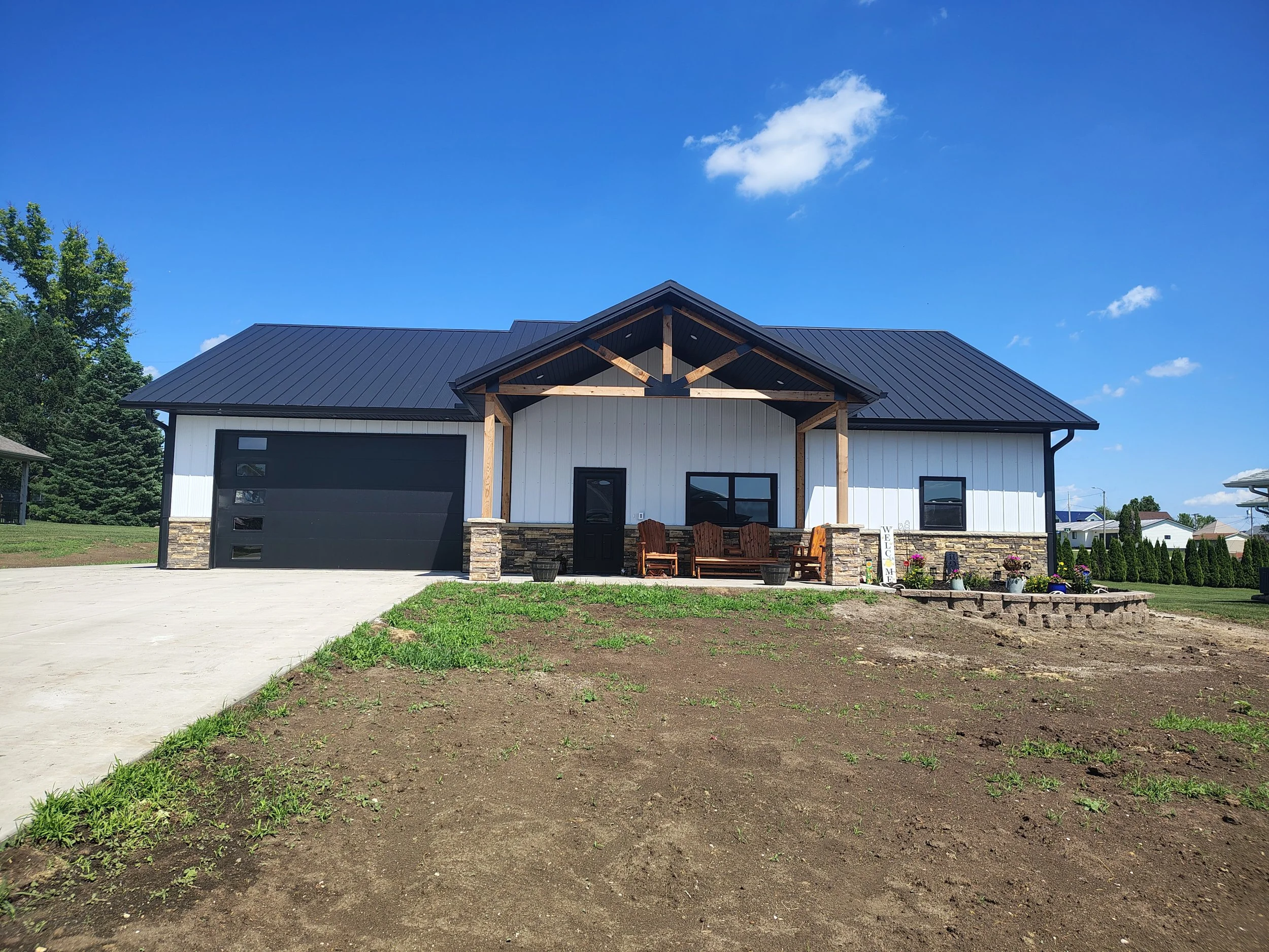 A modern house with a metal roof, white siding, a black garage door, stone accents, a front porch with wooden benches, and a landscaped front yard under a blue sky with some clouds.